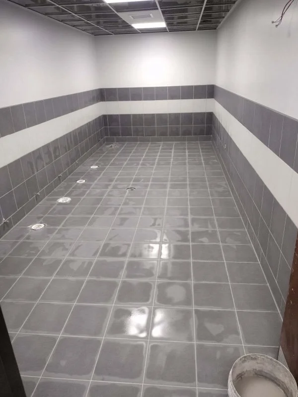 Empty tiled room with gray and white tiles, featuring floor drains and recessed lighting.