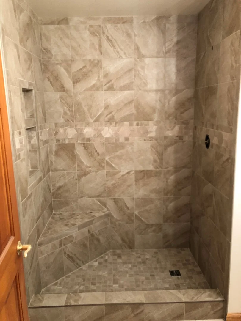 A tiled shower with beige and gray tiles, featuring a built-in bench and recessed shelf.