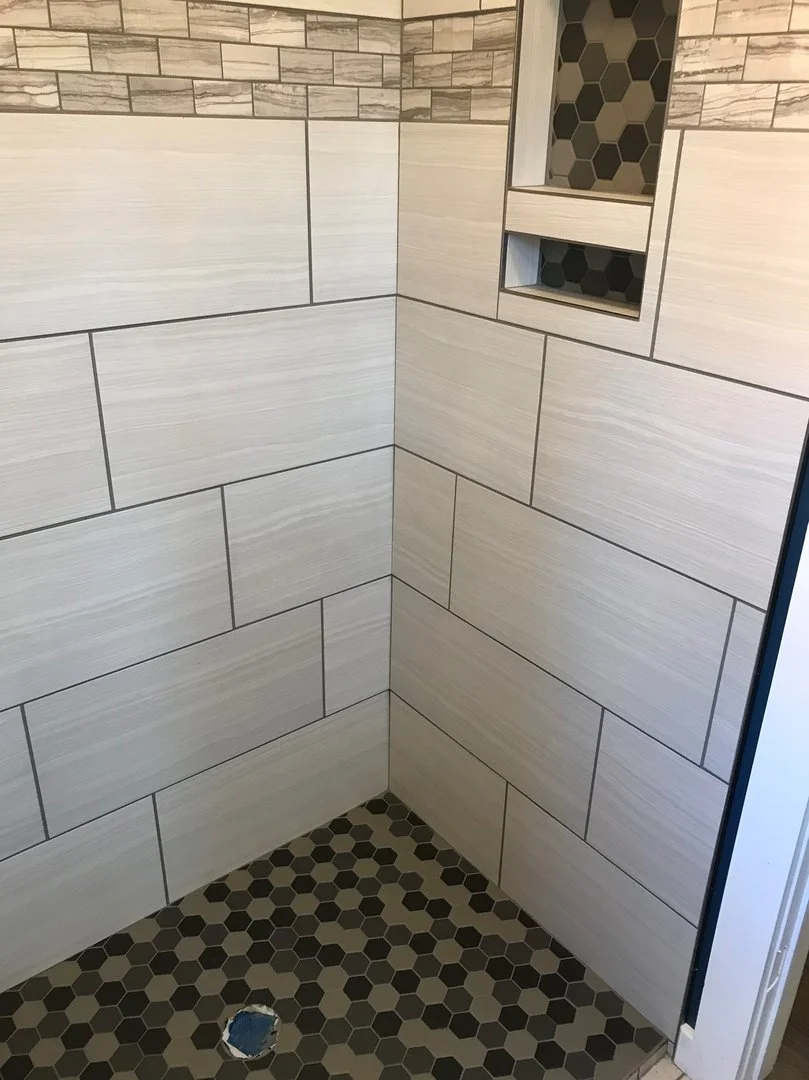 Bathroom shower corner with modern tile design, featuring light rectangular wall tiles, a decorative border, and hexagonal floor tiles.