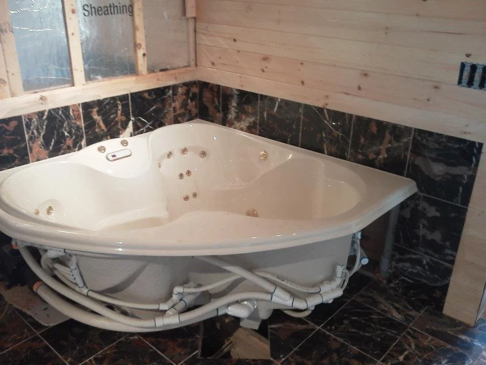Corner jacuzzi bathtub with tiled walls and exposed piping in a partially finished bathroom.