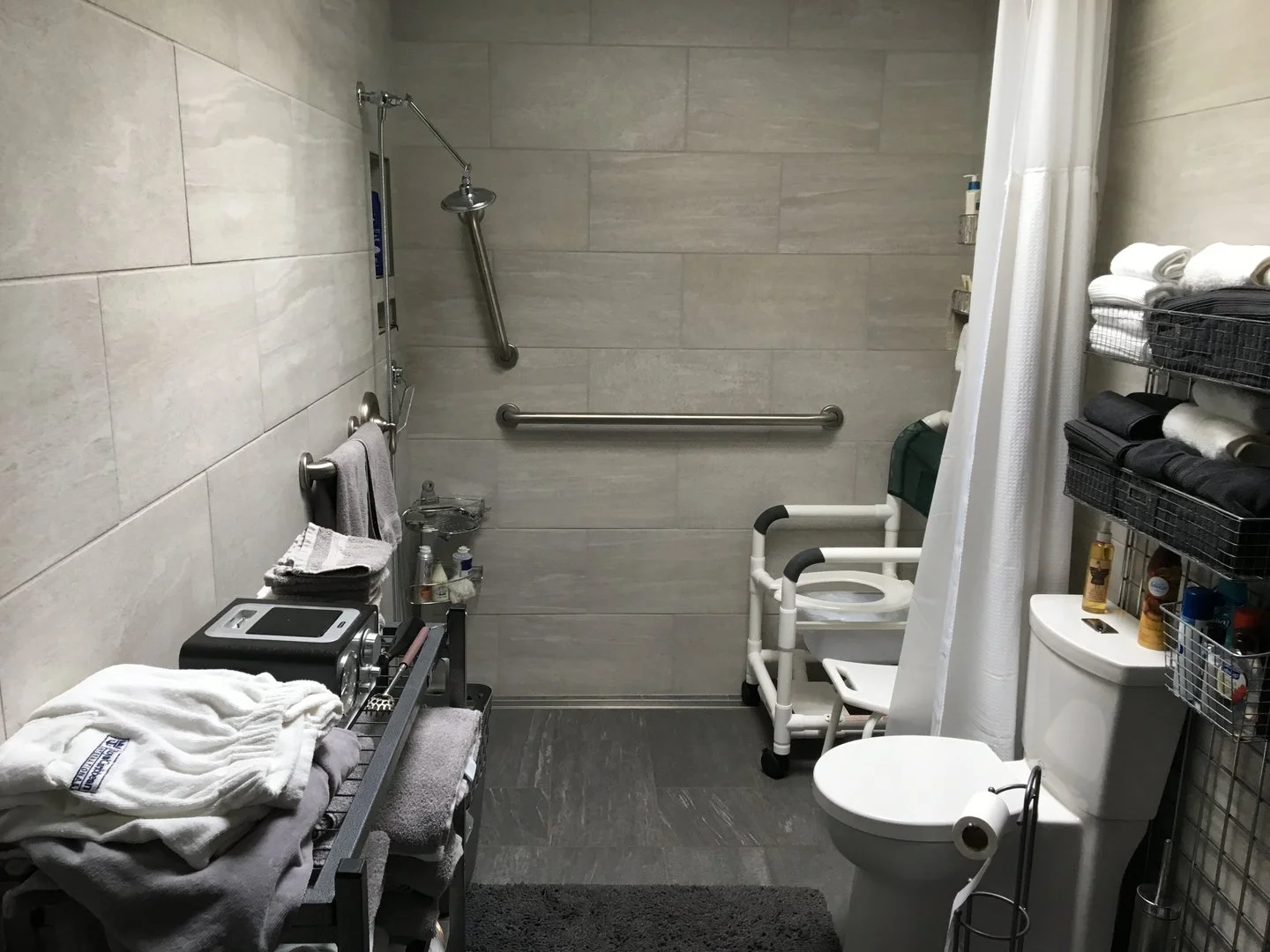 Accessible bathroom with shower and toilet, featuring grab bars, shower chair, towel rack, showerhead, storage shelves with towels, and hygiene products.
