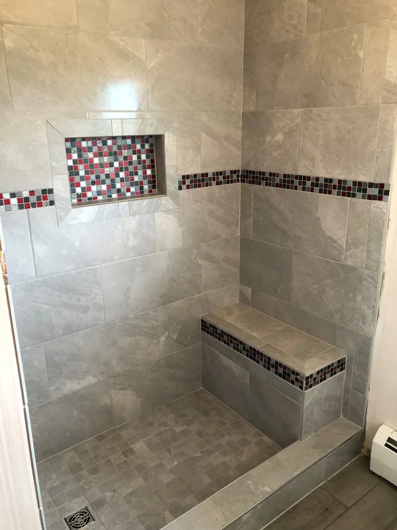 Modern shower with gray tiles, featuring a built-in bench and a recessed shelf. Decorative mosaic accents in red and black tiles adorn the wall and bench. A floor drain is visible on the shower floor.