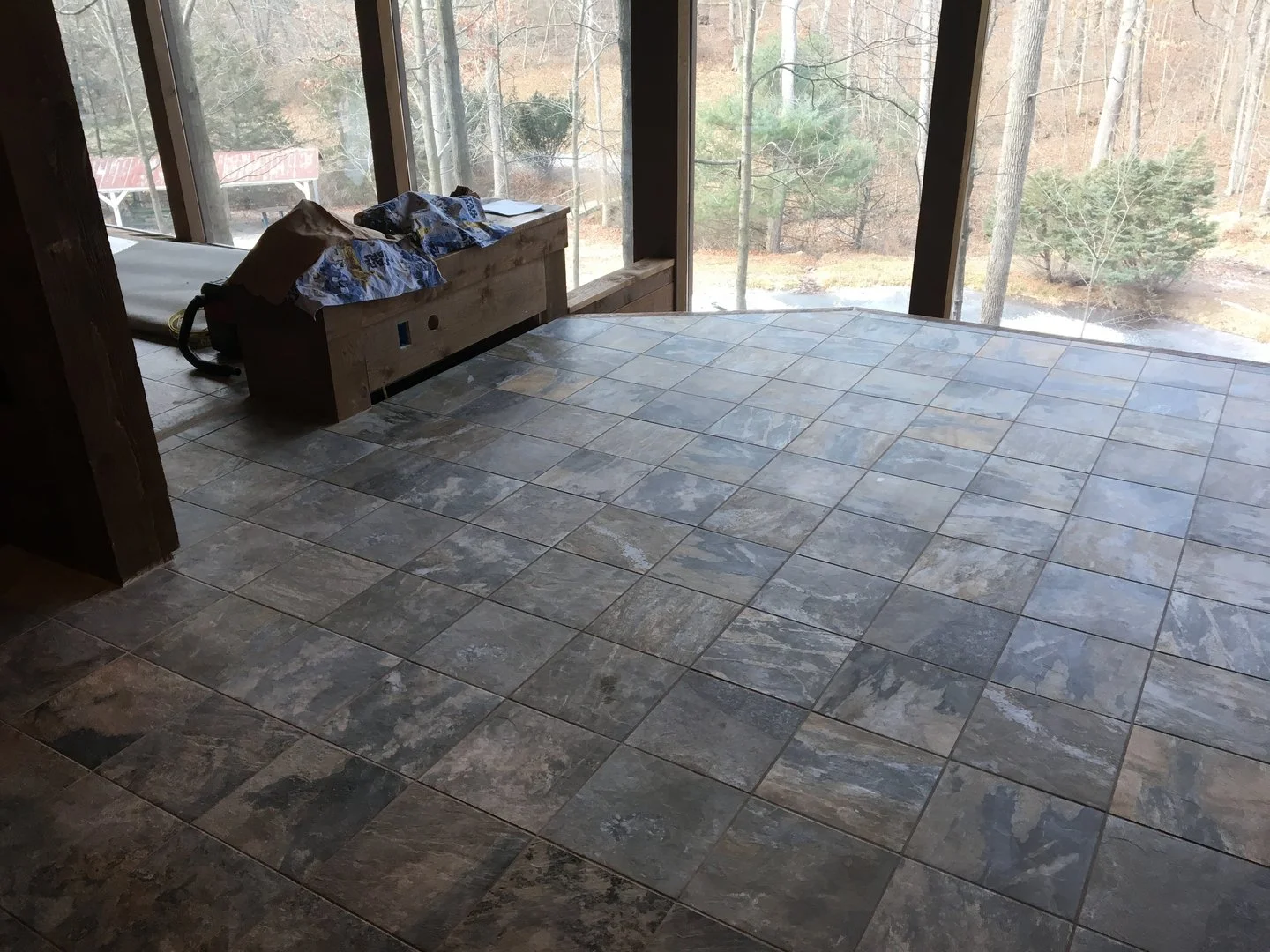 Interior room with stone tile flooring, large windows, and a wooded view outside. A wooden bench sits near the window with fabric draped over it.