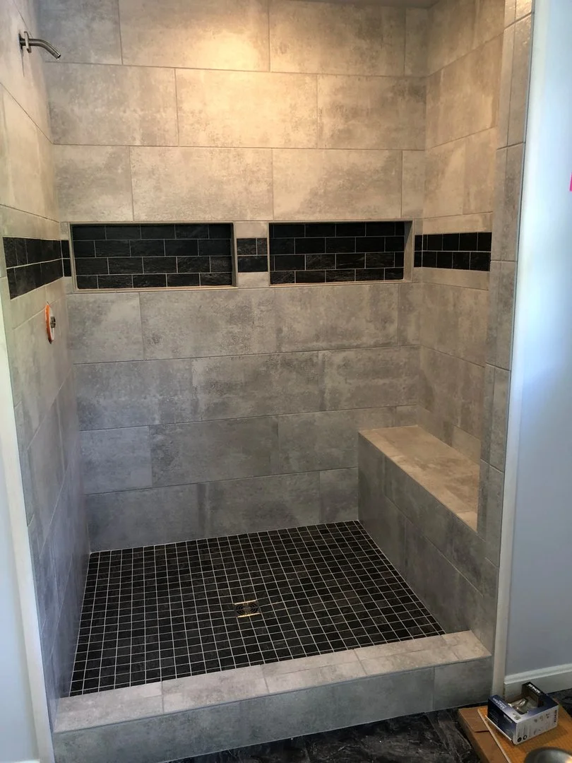 A modern shower with gray tiles and a built-in bench. The walls have a horizontal strip of darker tiles for contrast. The floor is tiled with small dark square tiles, and there is a recessed shelf area.