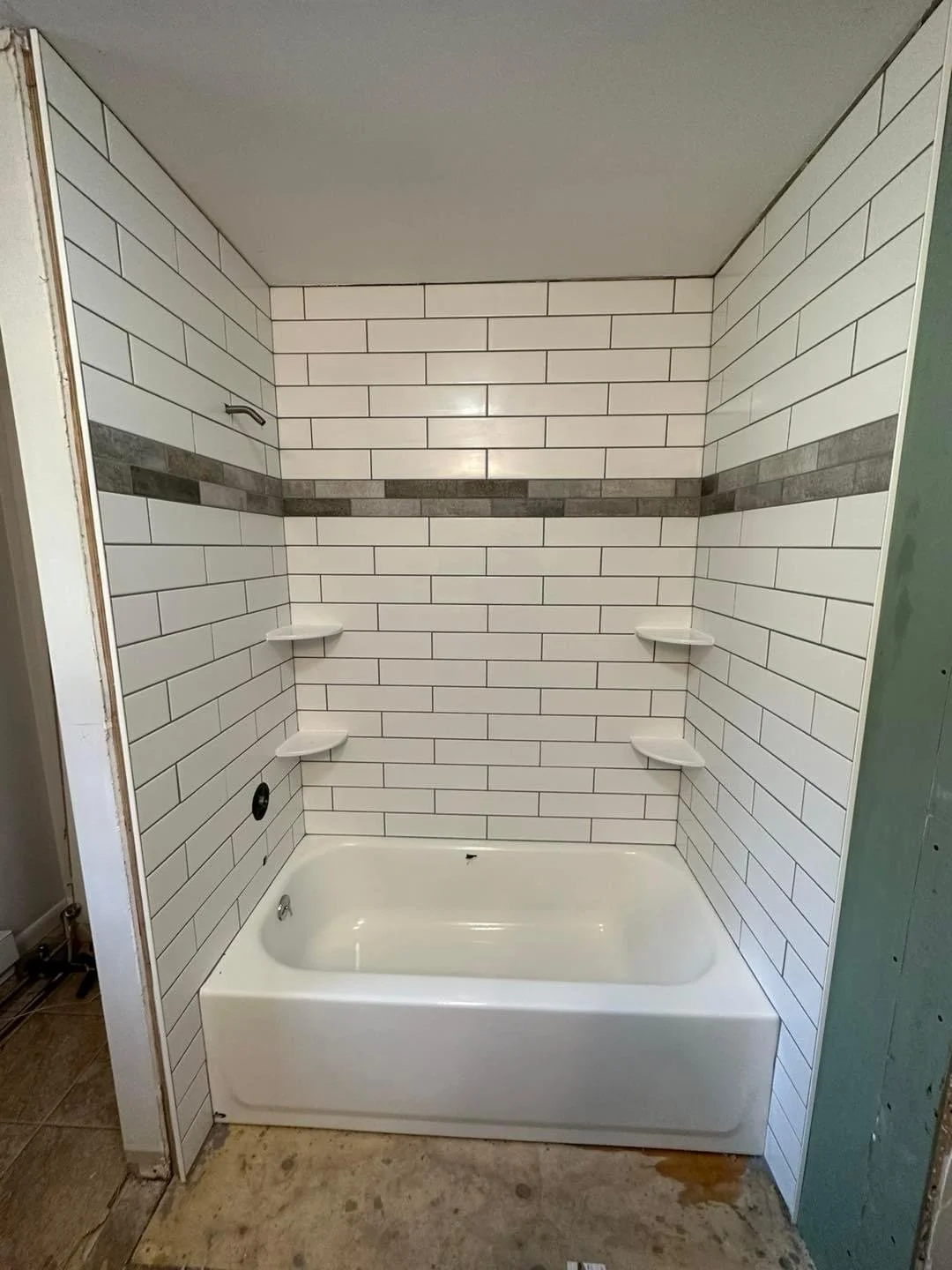 Newly installed bathtub with white subway tile walls, featuring a horizontal gray accent stripe and three corner shelves, in a bathroom under renovation.
