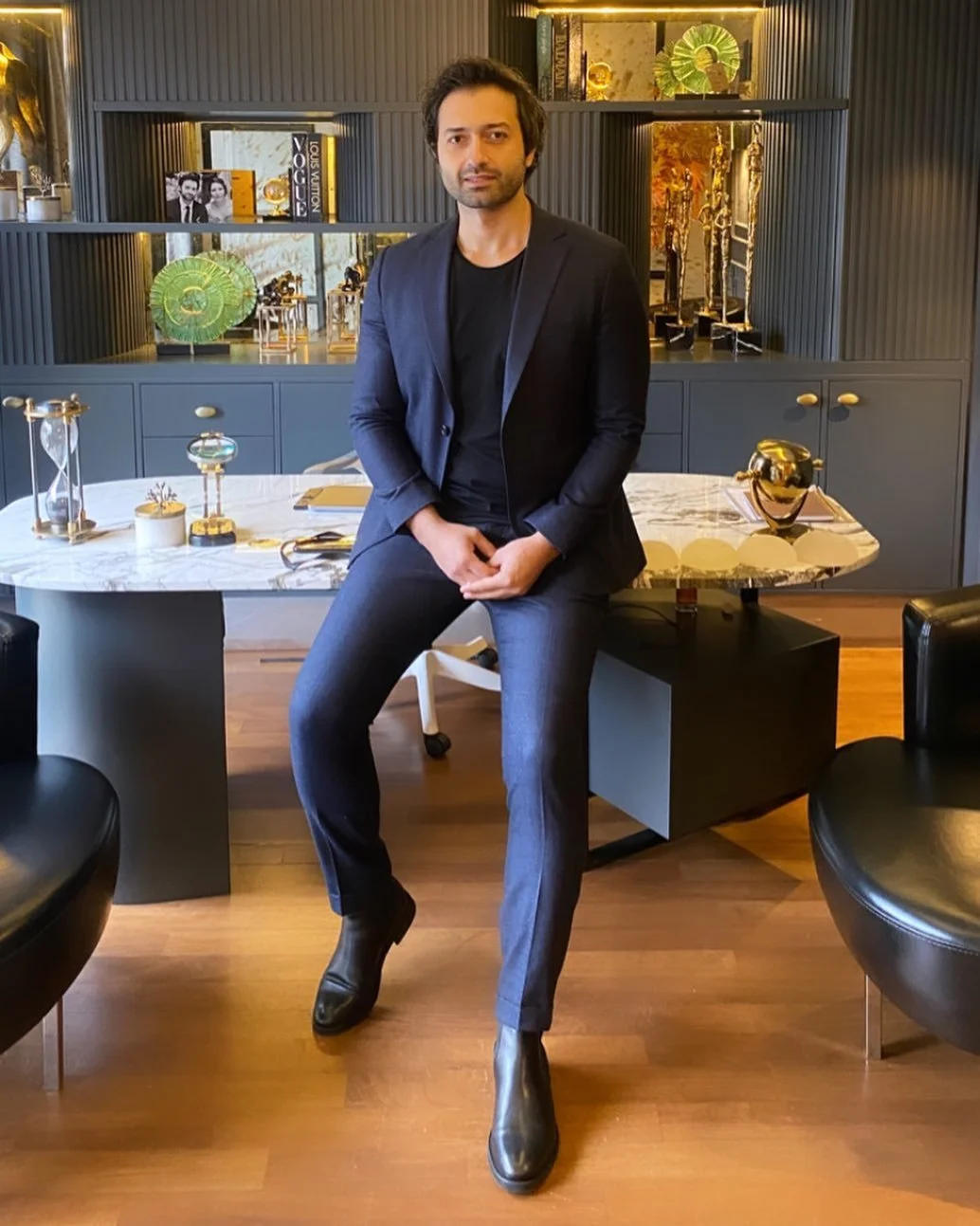 A man in a dark suit sits on the edge of an office desk. The room is stylish, with decorative items and shelves in the background, including trophies and books. The floor is wooden, and there are black chairs nearby.
