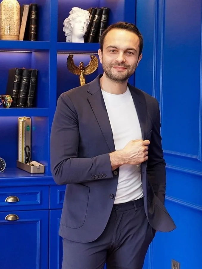 Man in a blue suit standing in front of a blue bookshelf with decorative items and books.