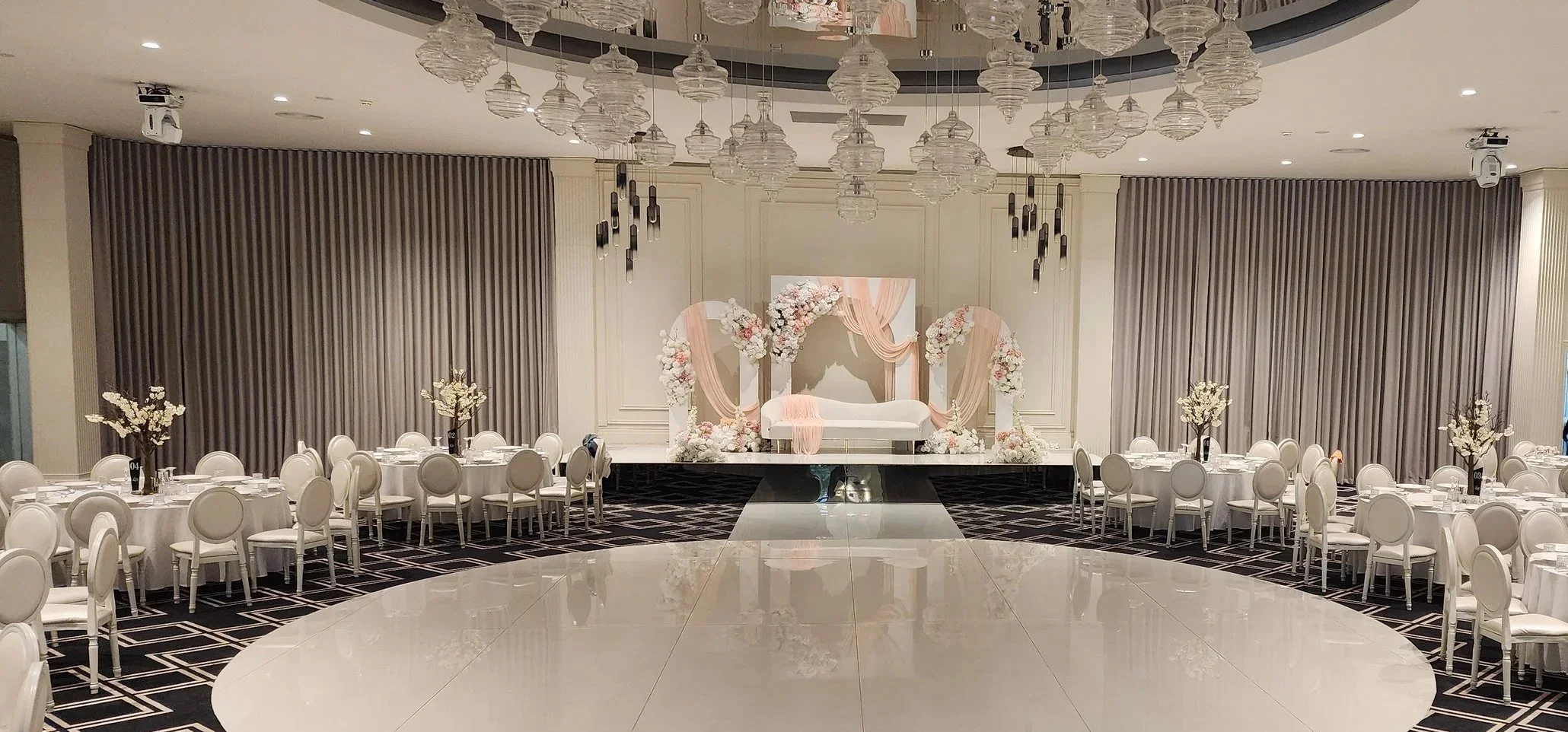 Elegant wedding decoration setup with white floral arch, draped fabric backdrop, and a white and gold bench. The foreground has a dining table with gold-rimmed plates, white napkins, and wine glasses.