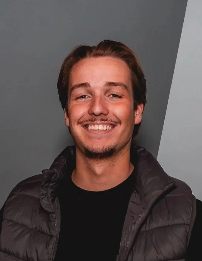 Een glimlachende jonge man met bruin haar, een lichte baard en een zwarte jas.