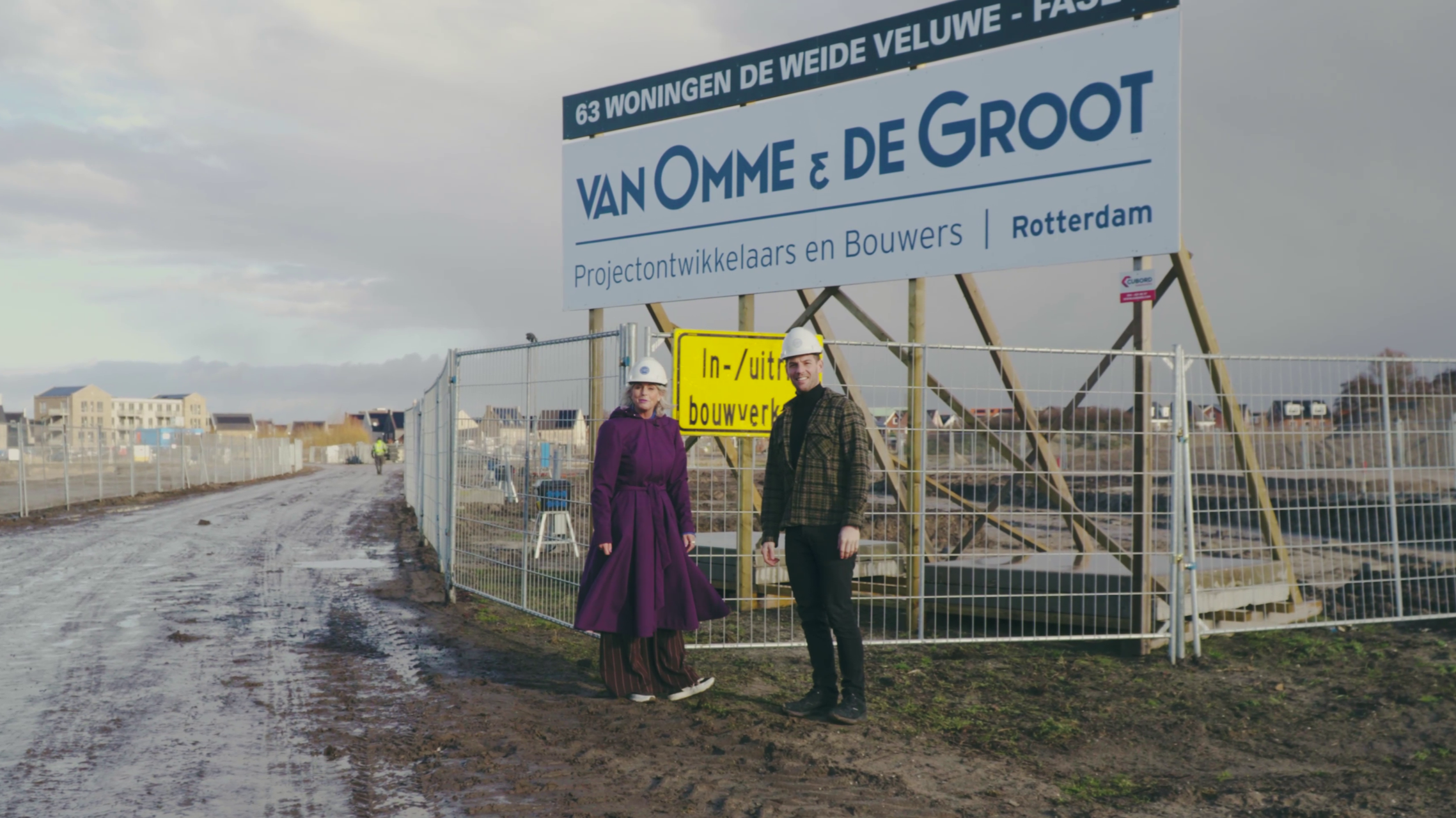 Twee personen met veiligheidhelmen voor een bouwplaats onder een groot bord met teksten over woningbouw in Rotterdam.