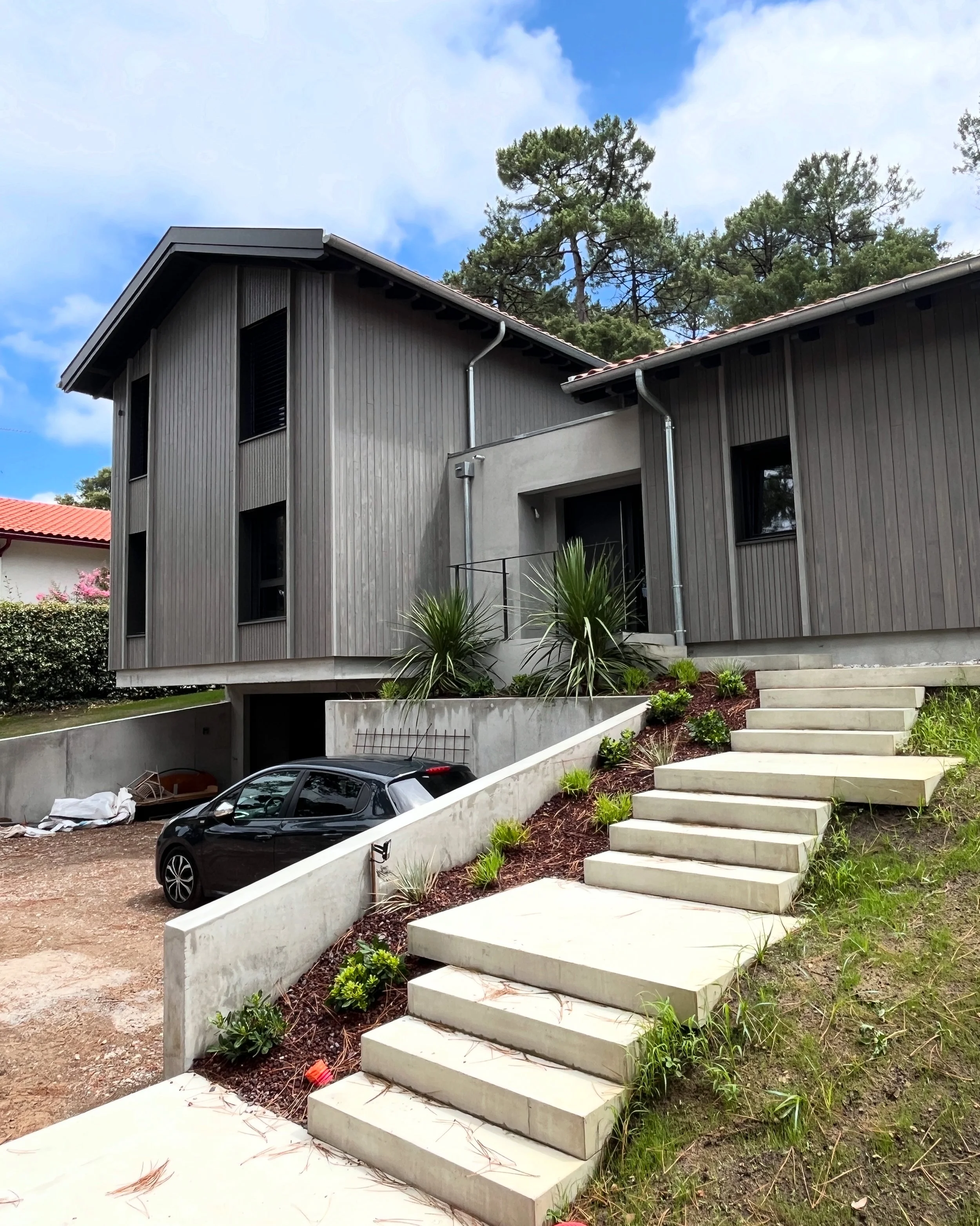Maison moderne avec escalier en béton menant au porche, voiture noire garée en dessous, végétation autour, ciel bleu avec quelques nuages.