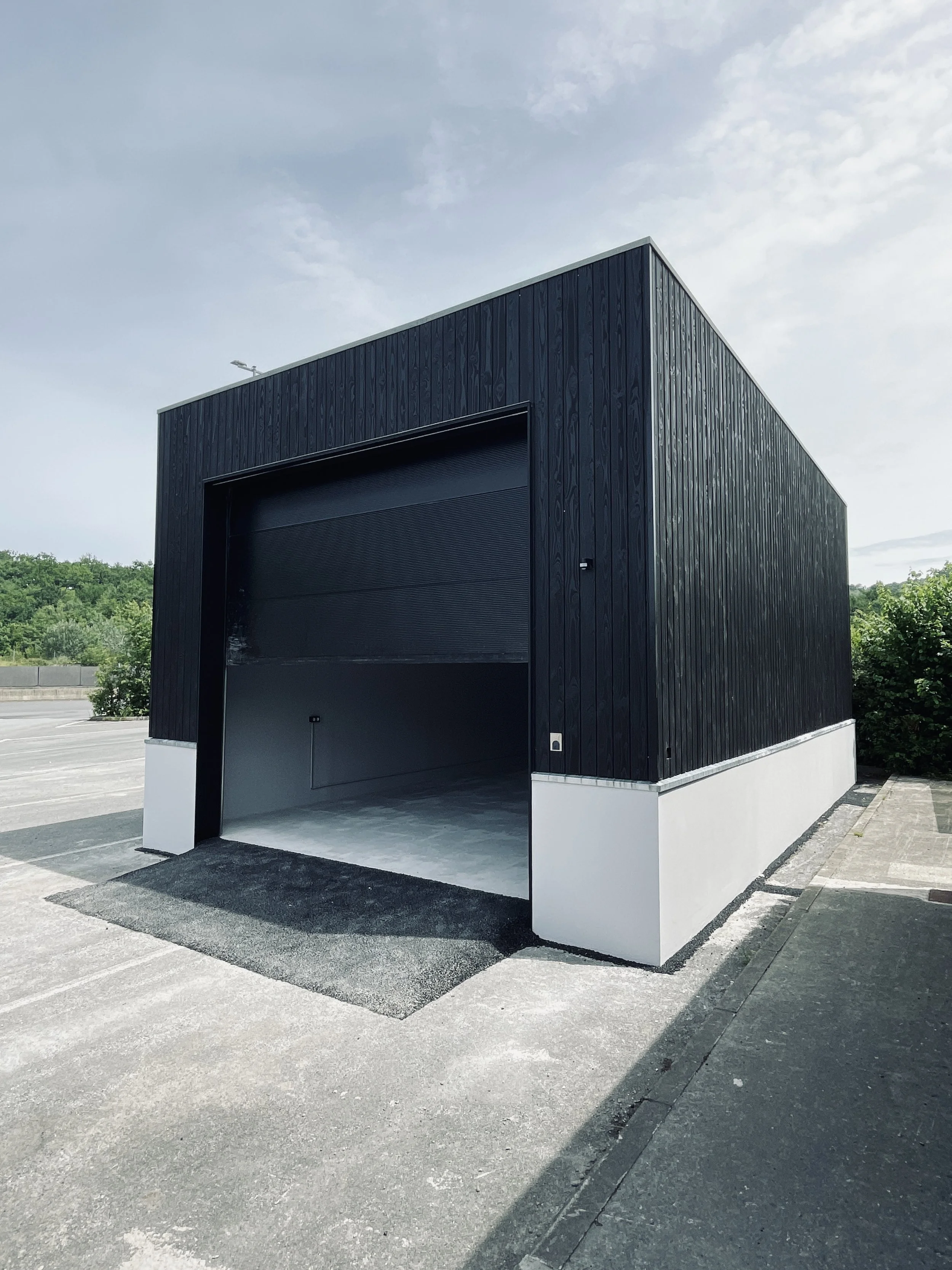 Un bâtiment noir moderne avec un grand garage en position ouverte, situé dans un parking extérieur.
