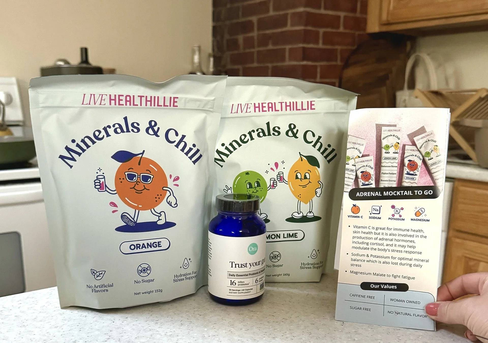 Various health supplement products on a kitchen counter, including two bags labeled 'Minerals & Chill' with animated citrus fruit characters, a small blue bottle labeled 'Trust your gut,' and a sheet of paper describing the product's benefits and values.