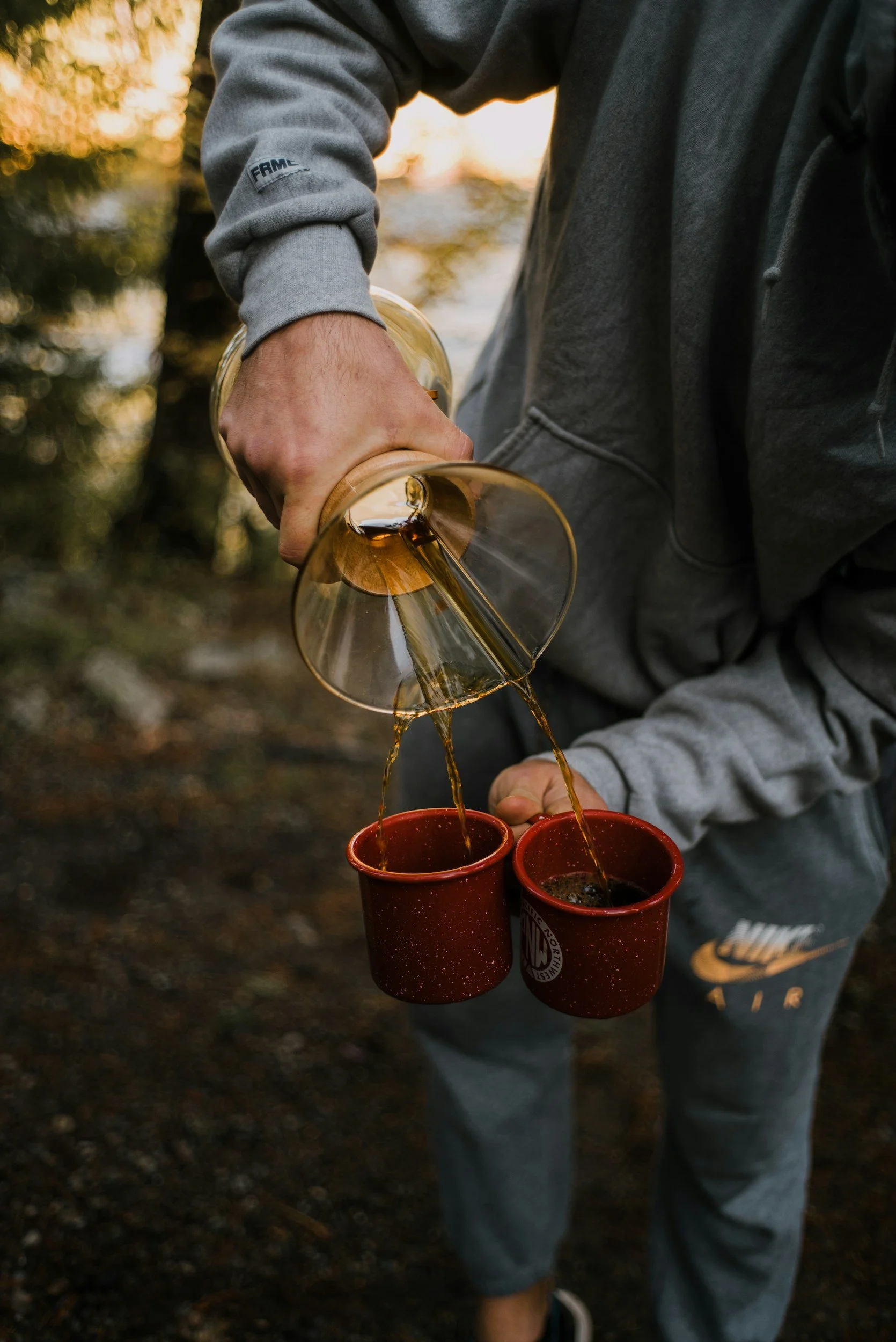 Person pouring coffee from a glass carafe into red cups outdoors during sunset.