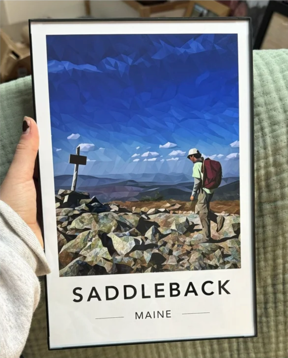 A framed poster of a hiker with a backpack walking past a wooden signpost on a rocky mountain summit under a blue sky with clouds, titled "Saddleback, Maine."