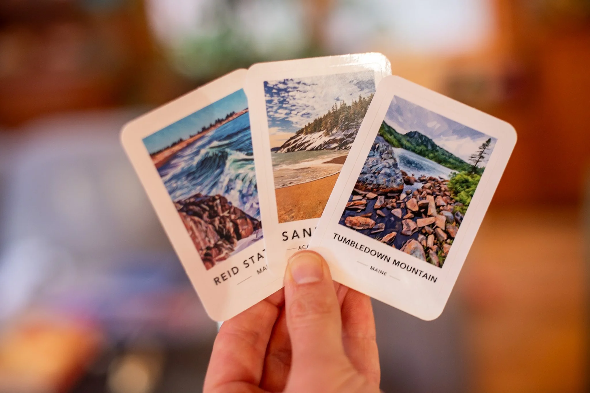 Hand holding three landscape-themed cards showing ocean waves, a mountain with clouds, and a river in a forest.