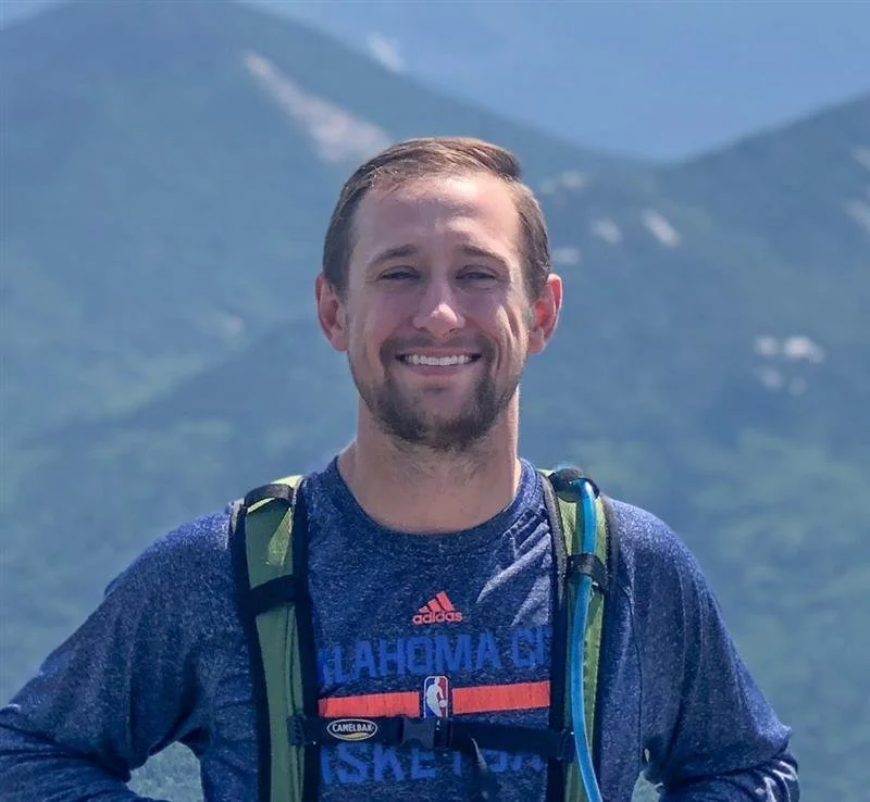 Man smiling outdoors with mountains and snowy peaks in the background, wearing a blue athletic shirt and a backpack.