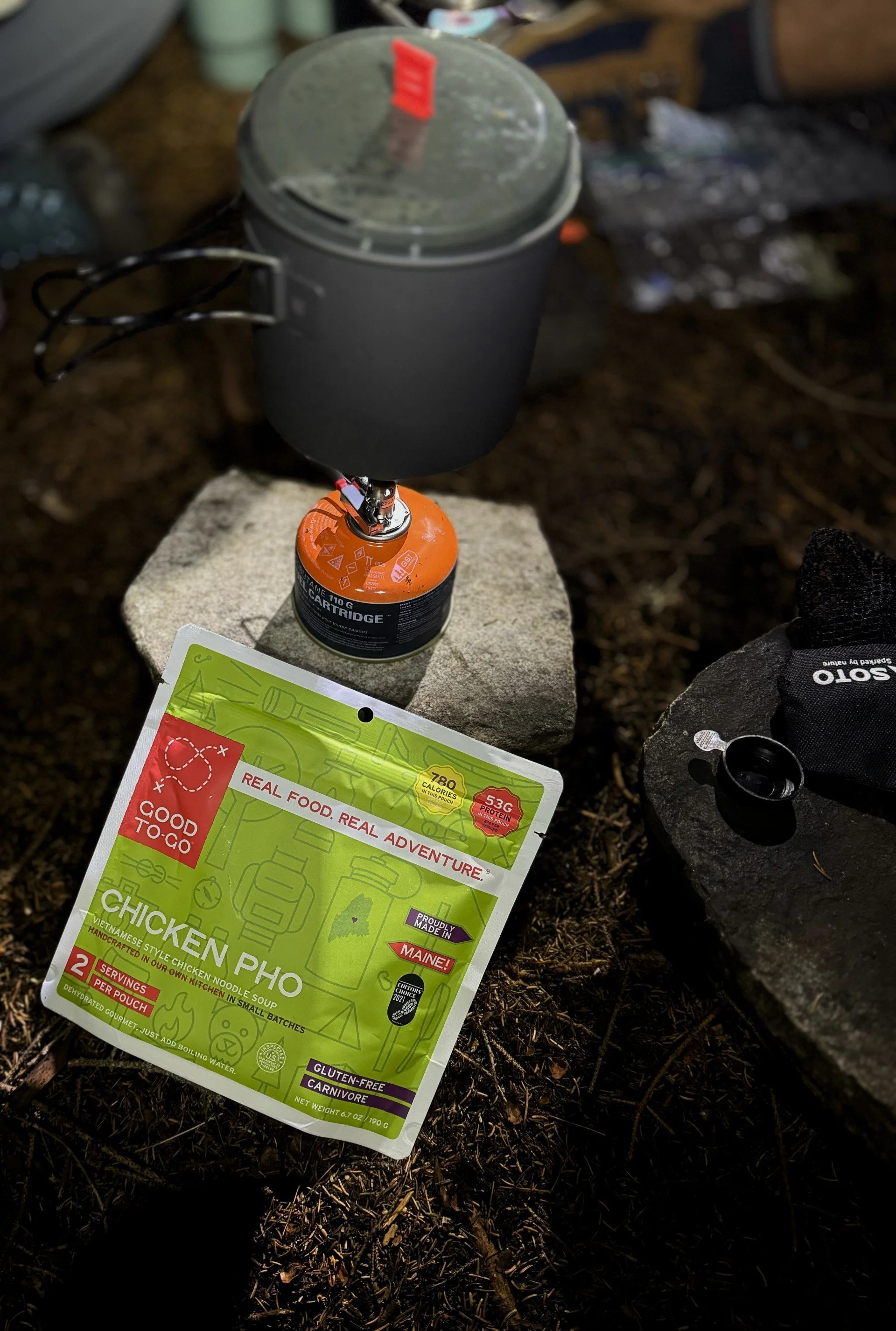 A camping stove with a canister of fuel, a black cooking pot, and a package of chicken pho noodle soup on a rock outside at night.