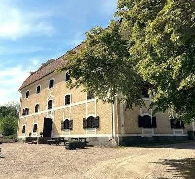 Historisk byggnad med gul väggfärg och svart tak, omgiven av träd med blommiga grenar, mot en klar blå himmel.