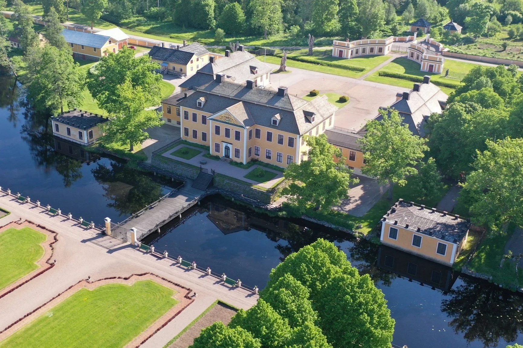 Lövstabruk - en av Sveriges bäst bevarade 1700-talsmiljöer
Luftbild av en stor, gul byggnad med många fönster och en mörk takkonstruktion, omgiven av gröna träd och gräsmattor, med en liten bro över en vattenarm och en parkeringsplats i närheten.