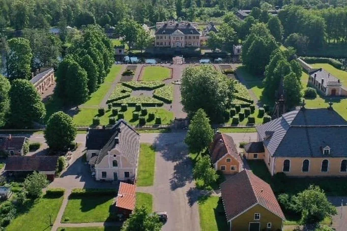 Flygbild av en trädgård med växthus, byggnader och gröna parker, omgiven av träd och ett stort föräldrahem i bakgrunden.