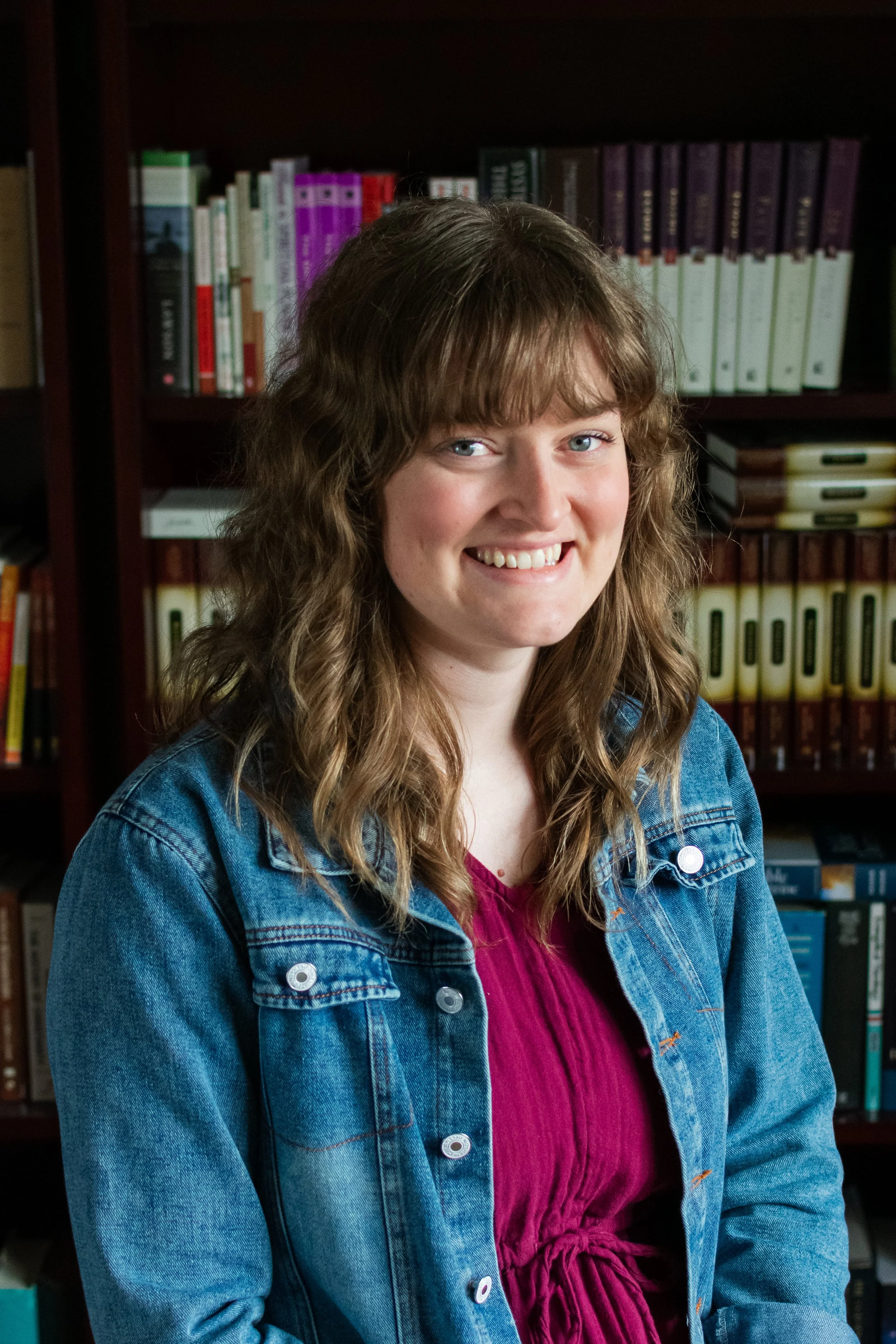 A young woman with curly light brown hair, blue eyes, and light skin, smiling, wearing a denim jacket over a magenta top, standing in front of a bookshelf filled with various books.