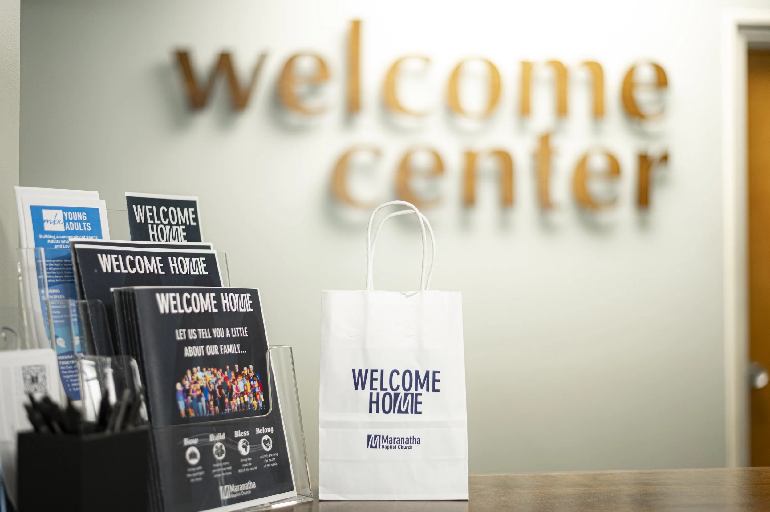 Welcome gift bag from Maranatha Baptist Church with a sign that says 'Welcome Home' in the foreground. Brochures and pens on a table.