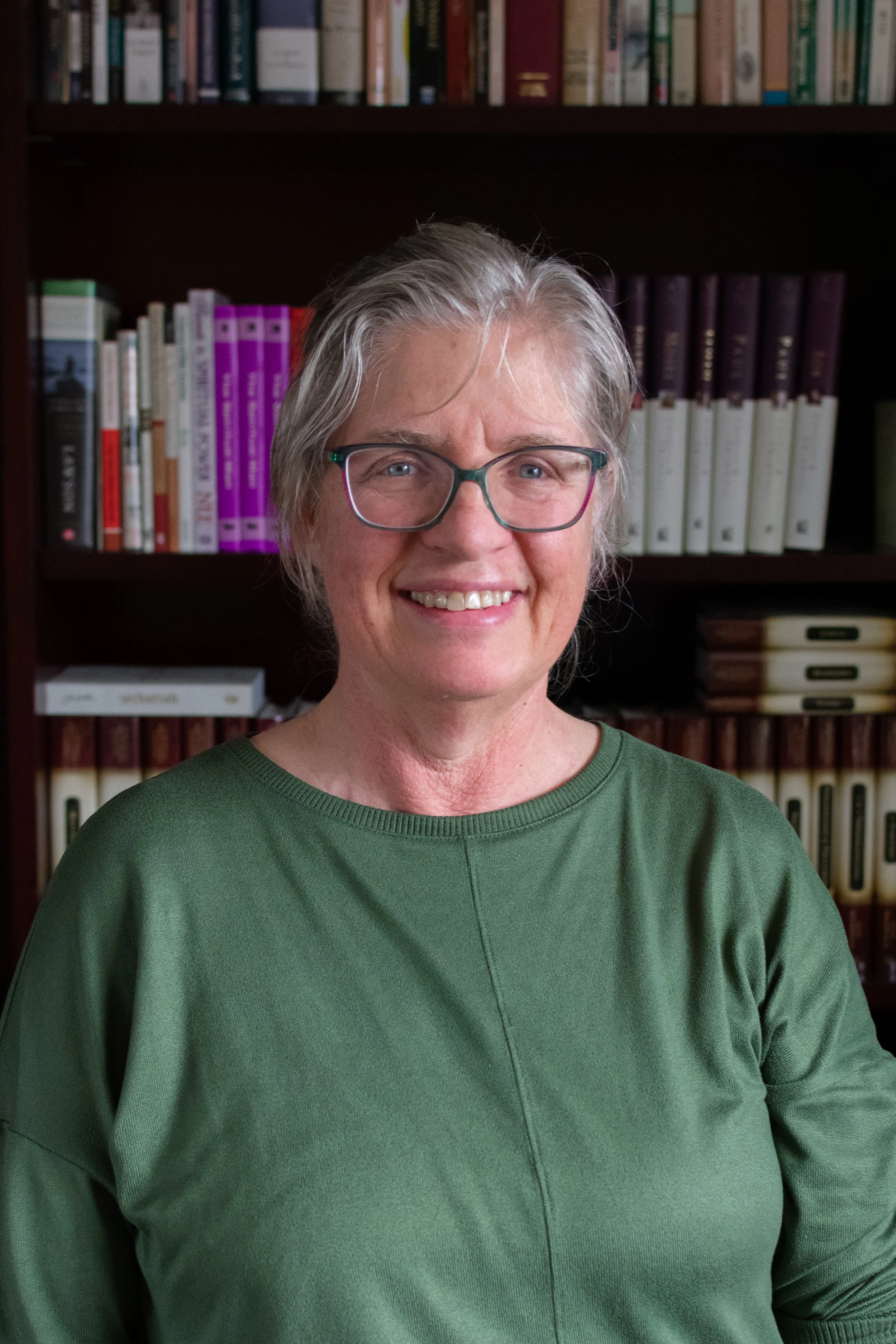 Photo of a smiling woman with gray hair and glasses, wearing a green top, standing in front of a bookshelf filled with various books.