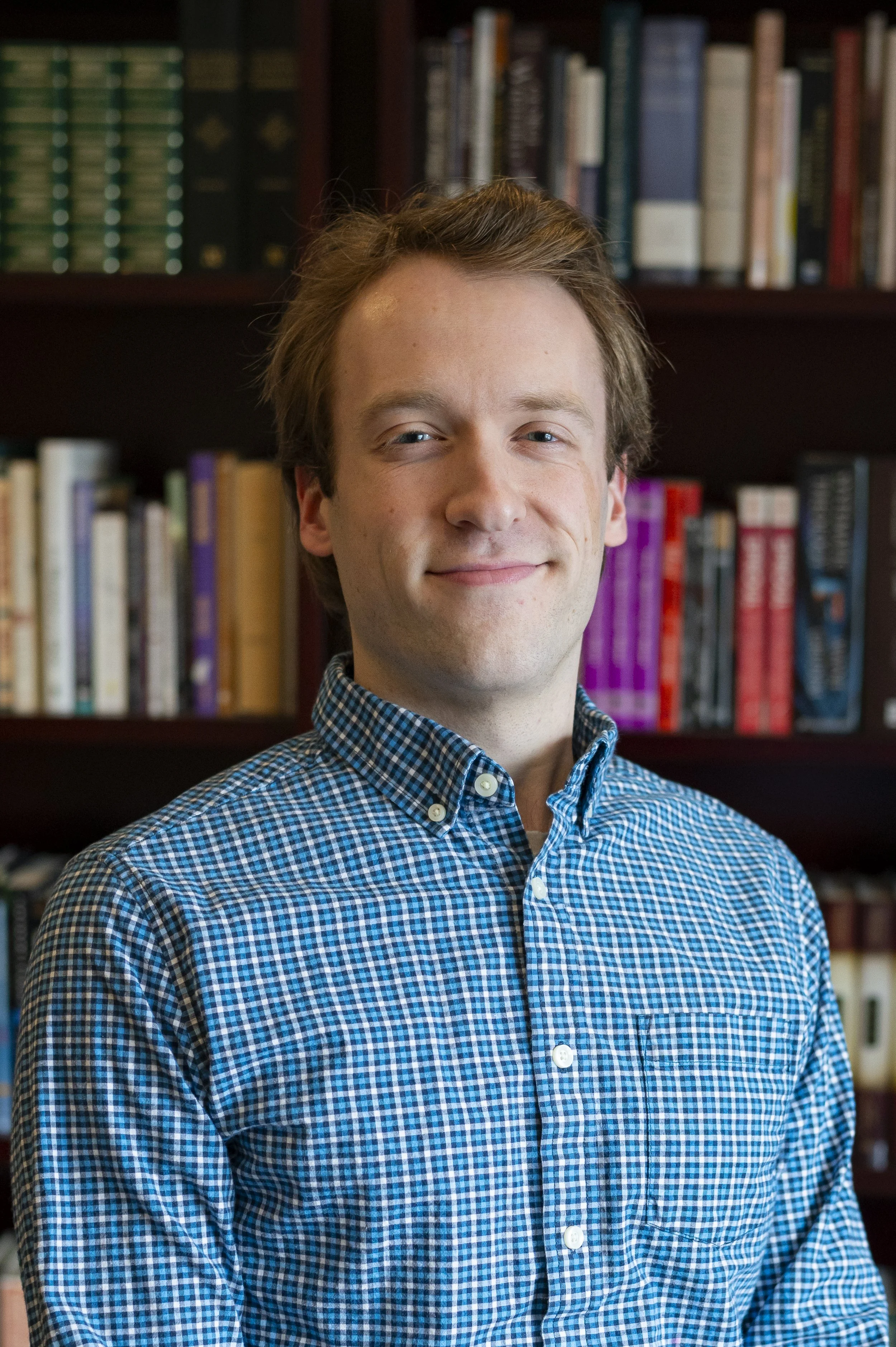 A young man with light skin, light brown hair, and blue eyes, wearing a blue and black checkered button-up shirt, standing in front of a bookshelf filled with books.