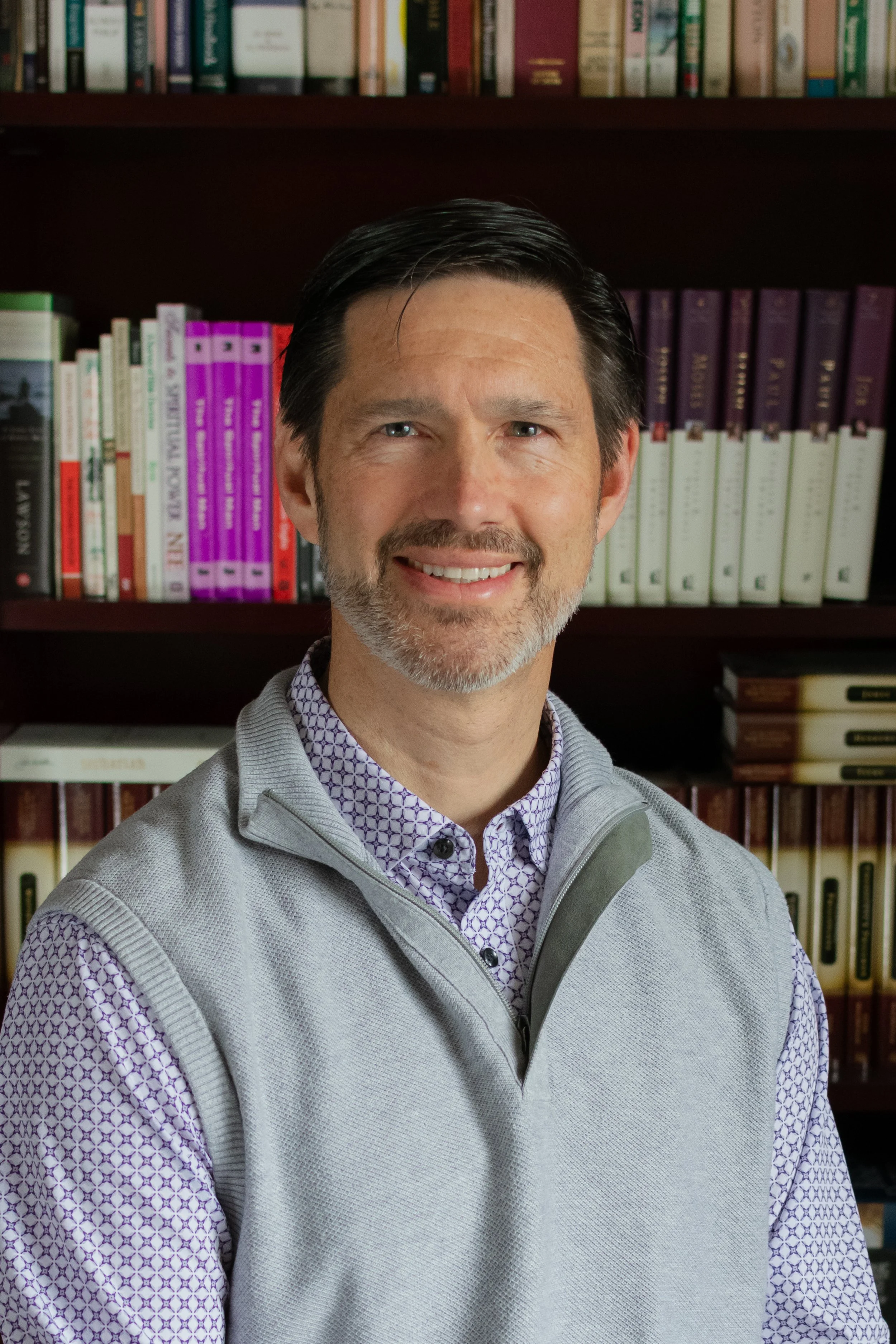 A portrait of a middle-aged man with dark hair and a beard, smiling, standing in front of a bookshelf filled with various colored books.