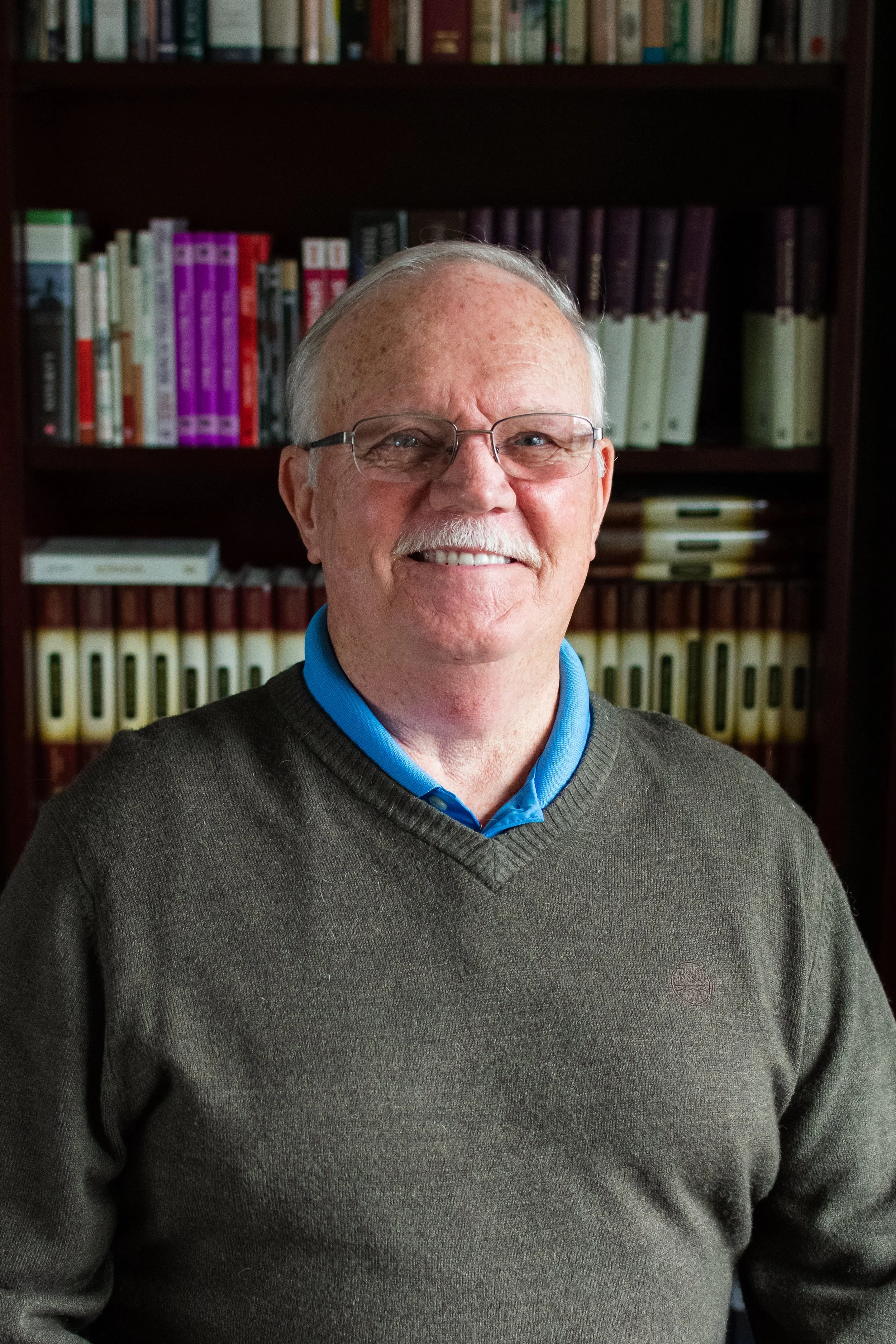 A smiling elderly man with glasses, wearing a blue collared shirt and a dark sweater, standing in front of a bookshelf filled with colorful books.