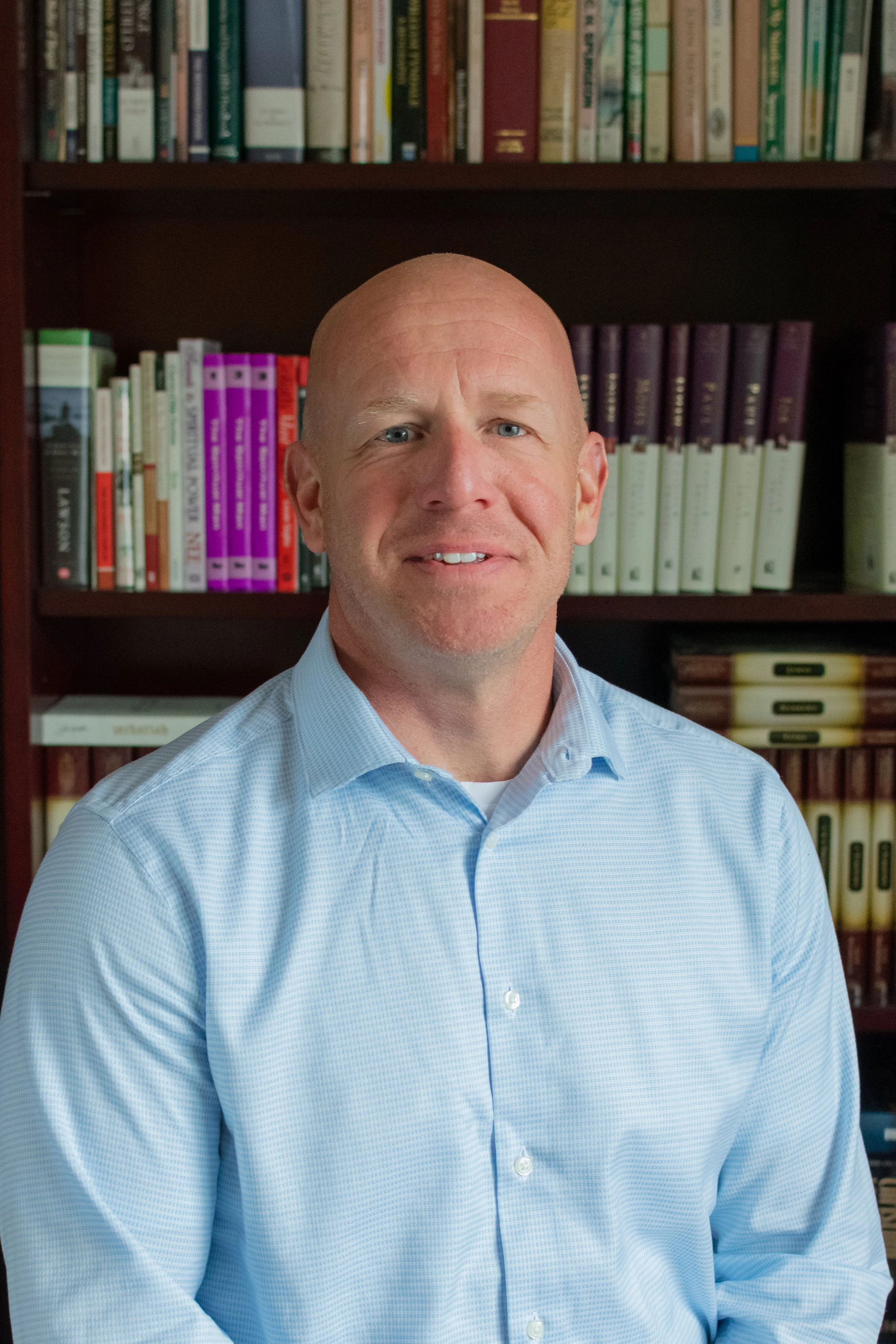 A man with a shaved head and blue eyes smiling in front of a bookshelf filled with various books.
