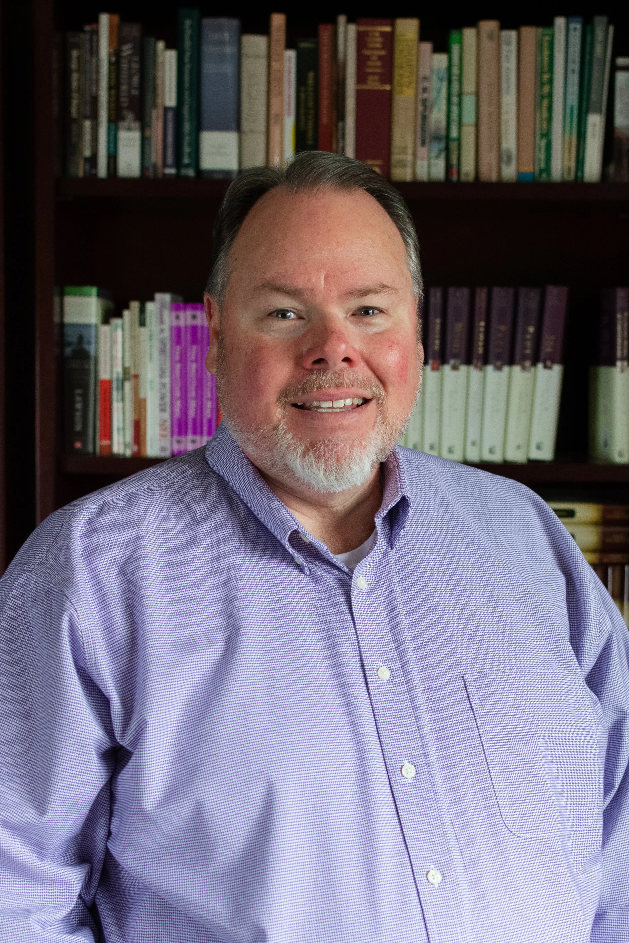 A man with gray hair and a beard smiling, wearing a purple checkered button-up shirt, in front of shelves filled with books.
