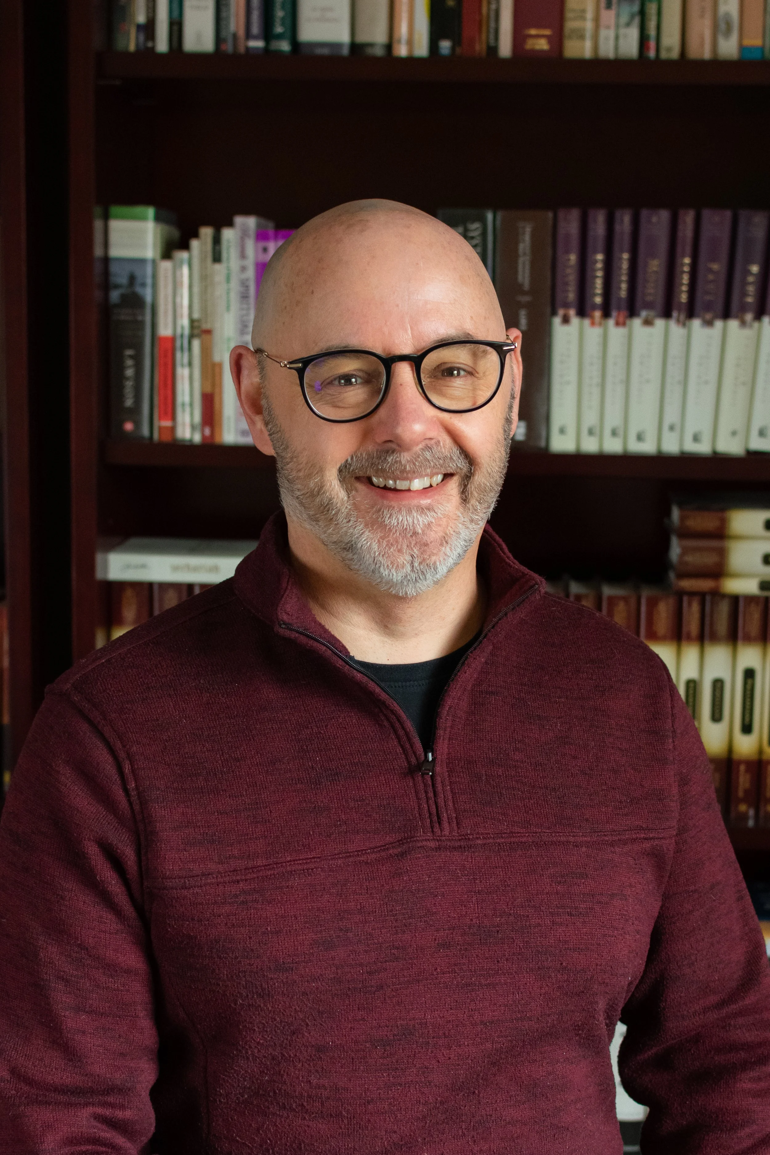 A smiling bald man with glasses, wearing a maroon zip-up sweater, standing in front of a bookshelf filled with books.