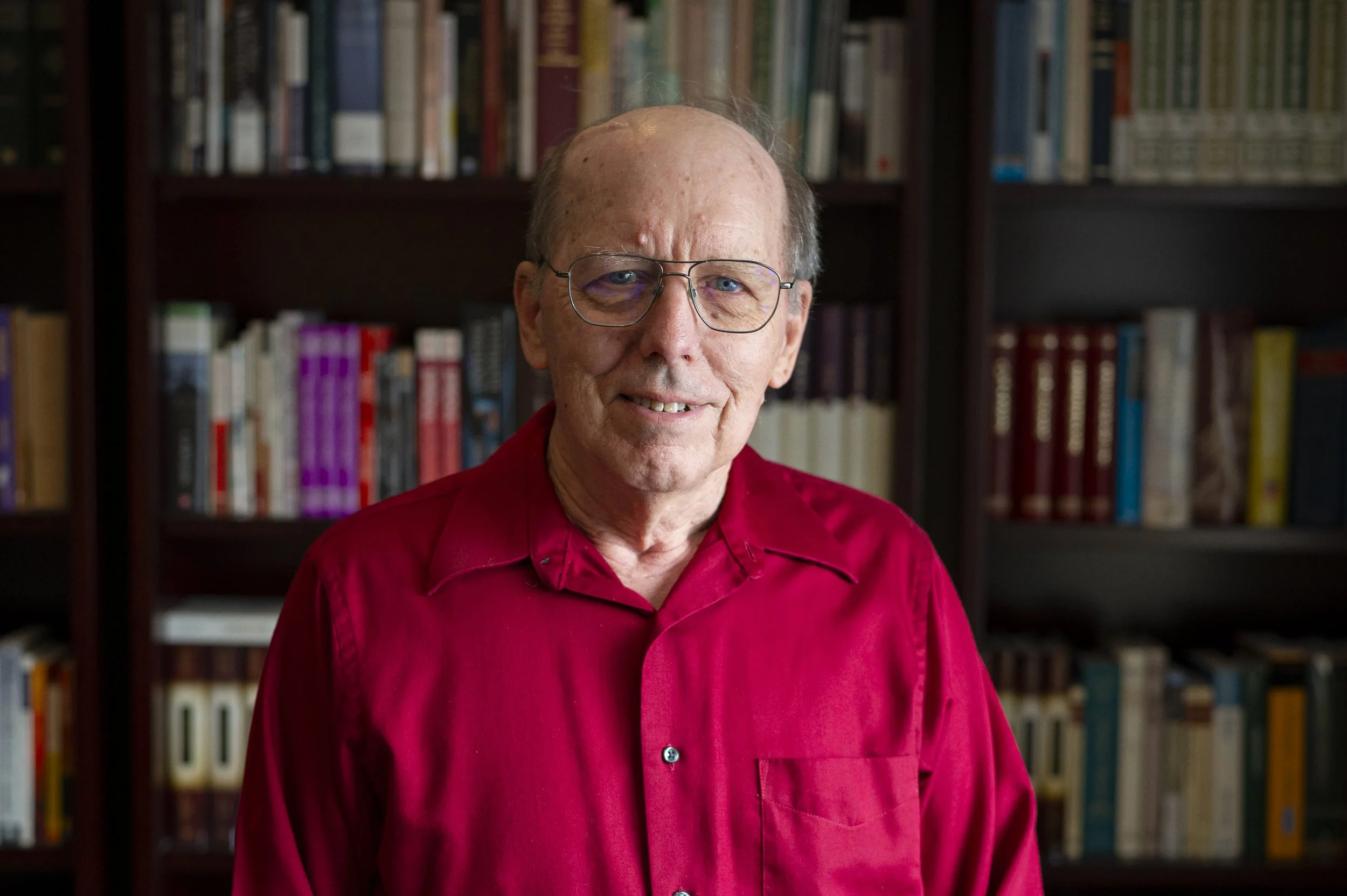 An elderly man with glasses and a red shirt standing in front of a bookshelf filled with books.