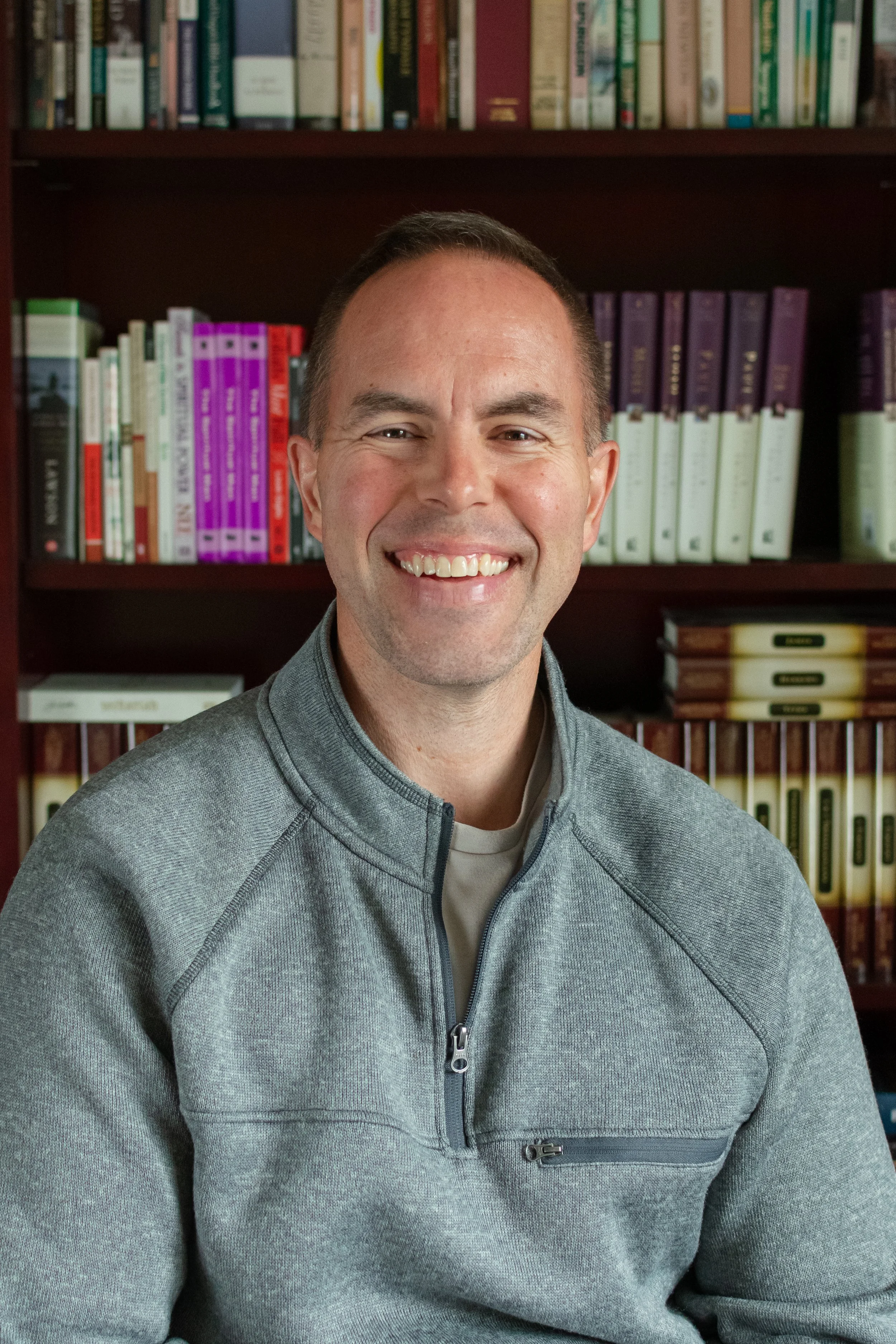 A smiling man with short brown hair wearing a gray zip-up jacket, standing in front of bookshelves filled with colorful books.
