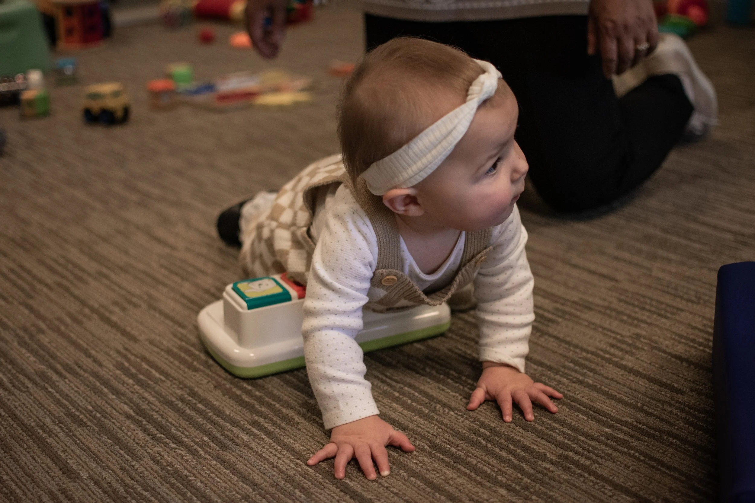 A young child with light hair and wearing a beige and white outfit, crawling on a striped carpet with a toy on their back, in a room with scattered toys and a person in sneakers nearby.