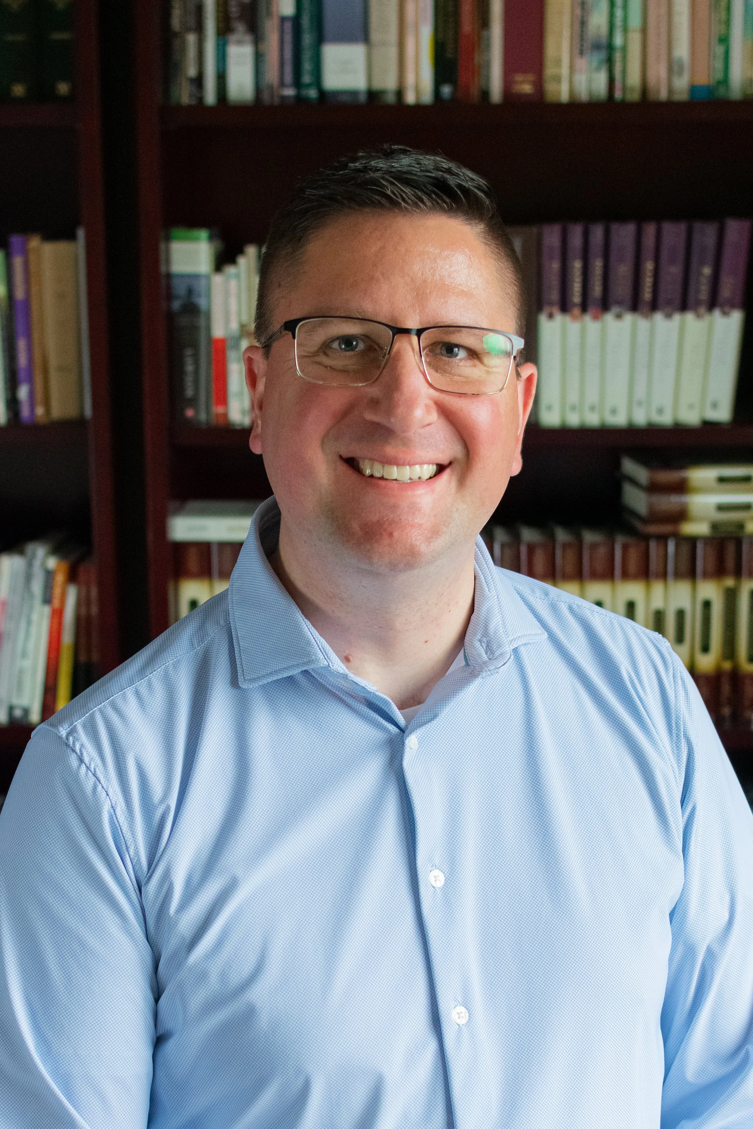 A man wearing glasses and a light blue collared shirt, smiling, standing in front of a bookshelf filled with books.