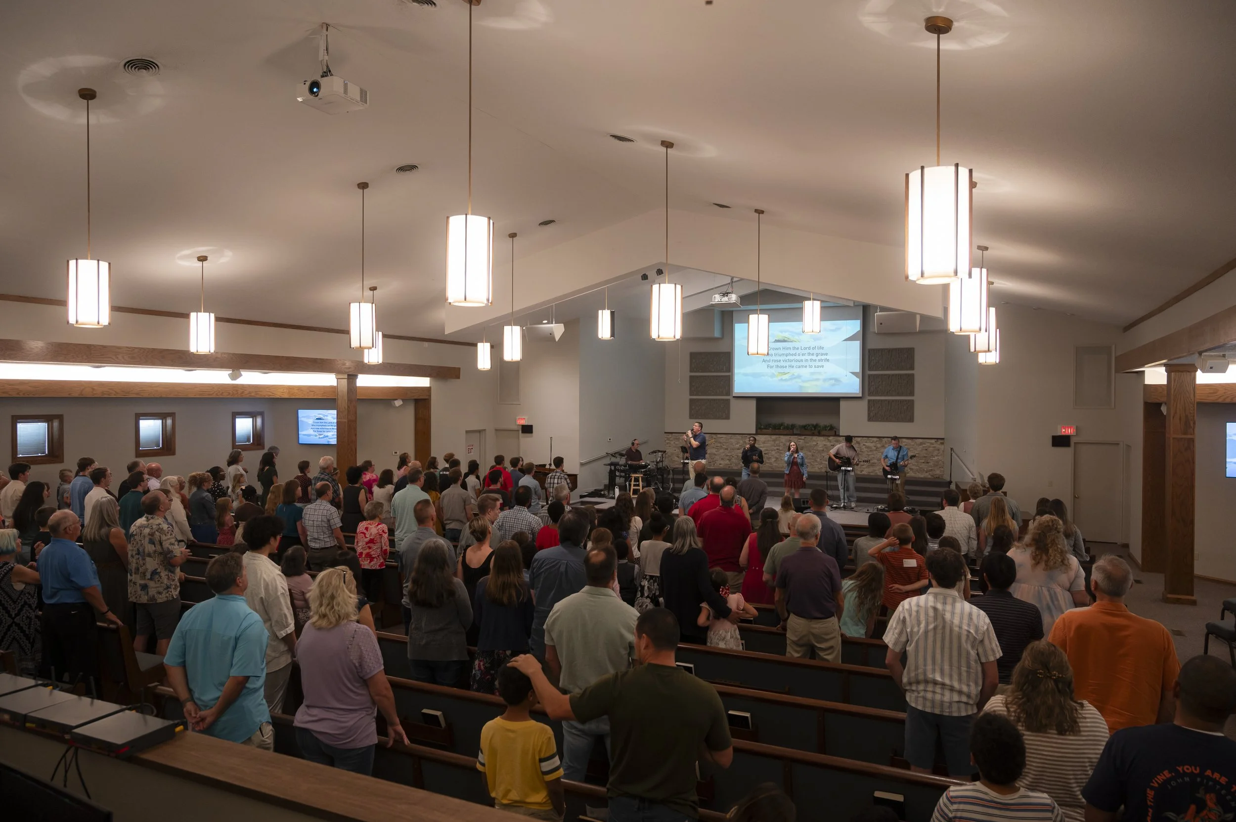 A large congregation gathered inside a church for a worship service or event, with a stage at the front where musicians and a person are singing, playing guitars, and leading the worship. There are multiple hanging lights on the ceiling and a projector displaying song lyrics or scripture on a screen behind the stage. The audience is standing, some with hands raised, participating in the service.