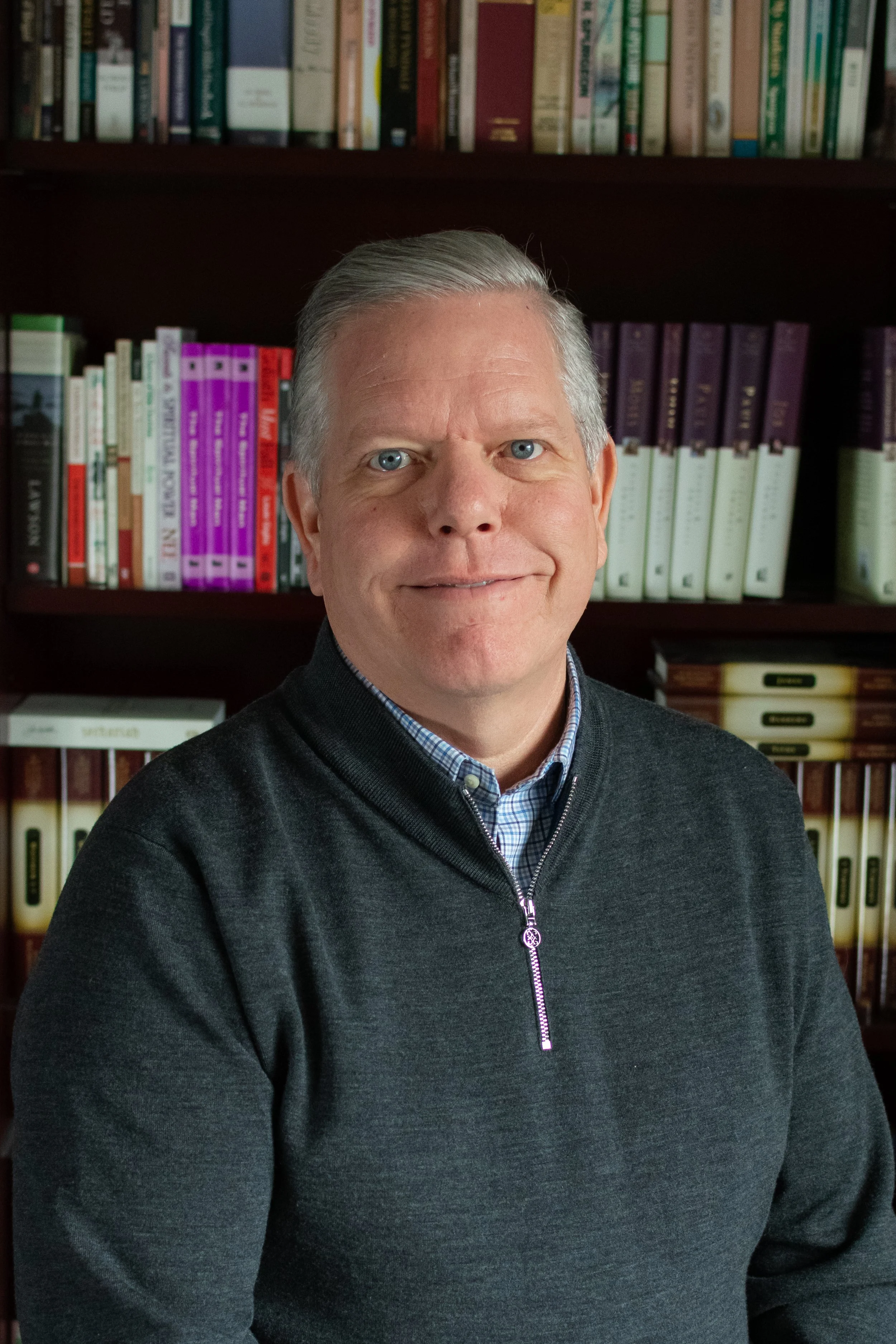 A middle-aged man with gray hair and blue eyes smiling, wearing a dark gray zip-up sweater over a blue checkered shirt, standing in front of a bookshelf filled with books.