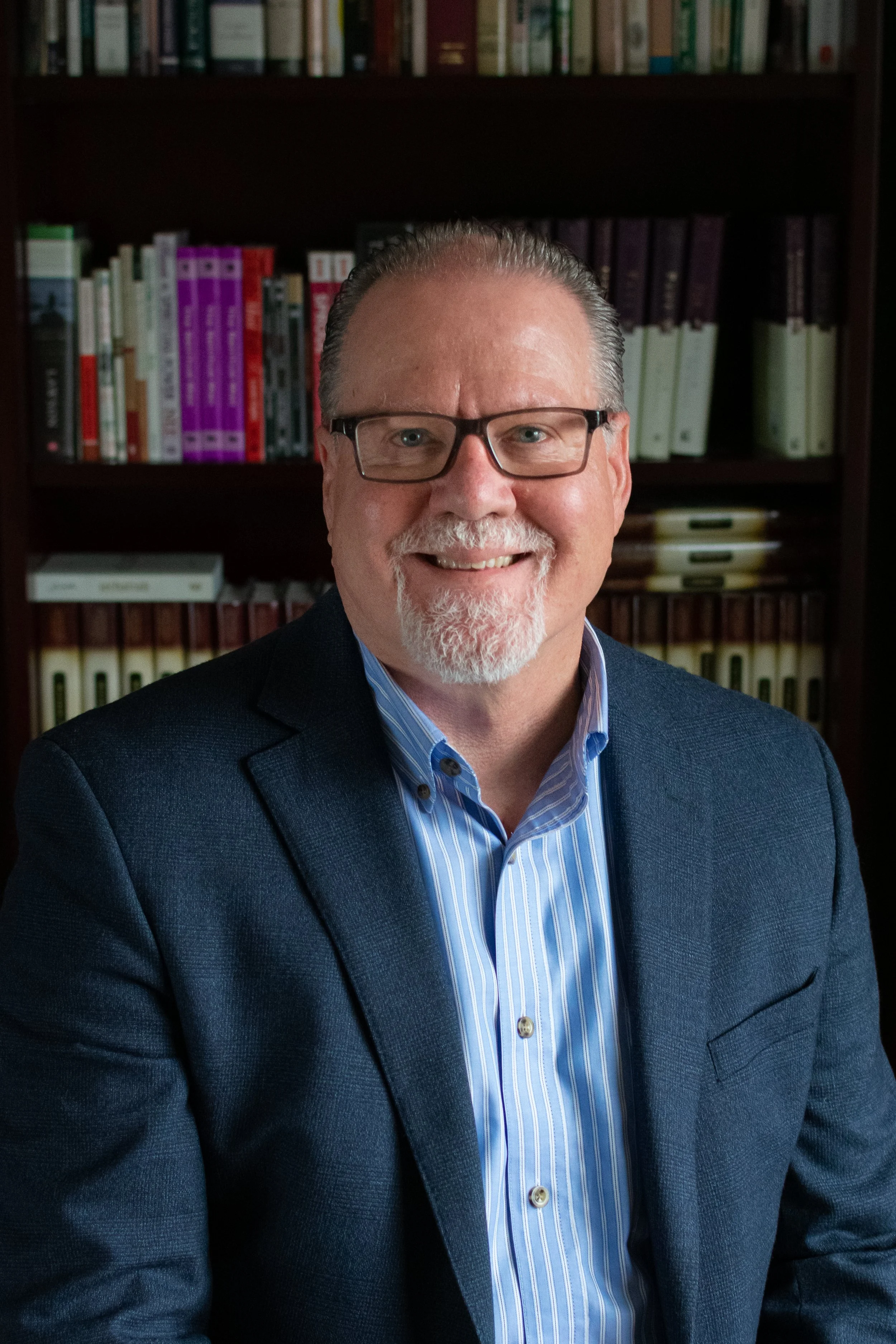 A middle-aged man with glasses and a beard in a suit jacket smiling, standing in front of a bookshelf filled with books.