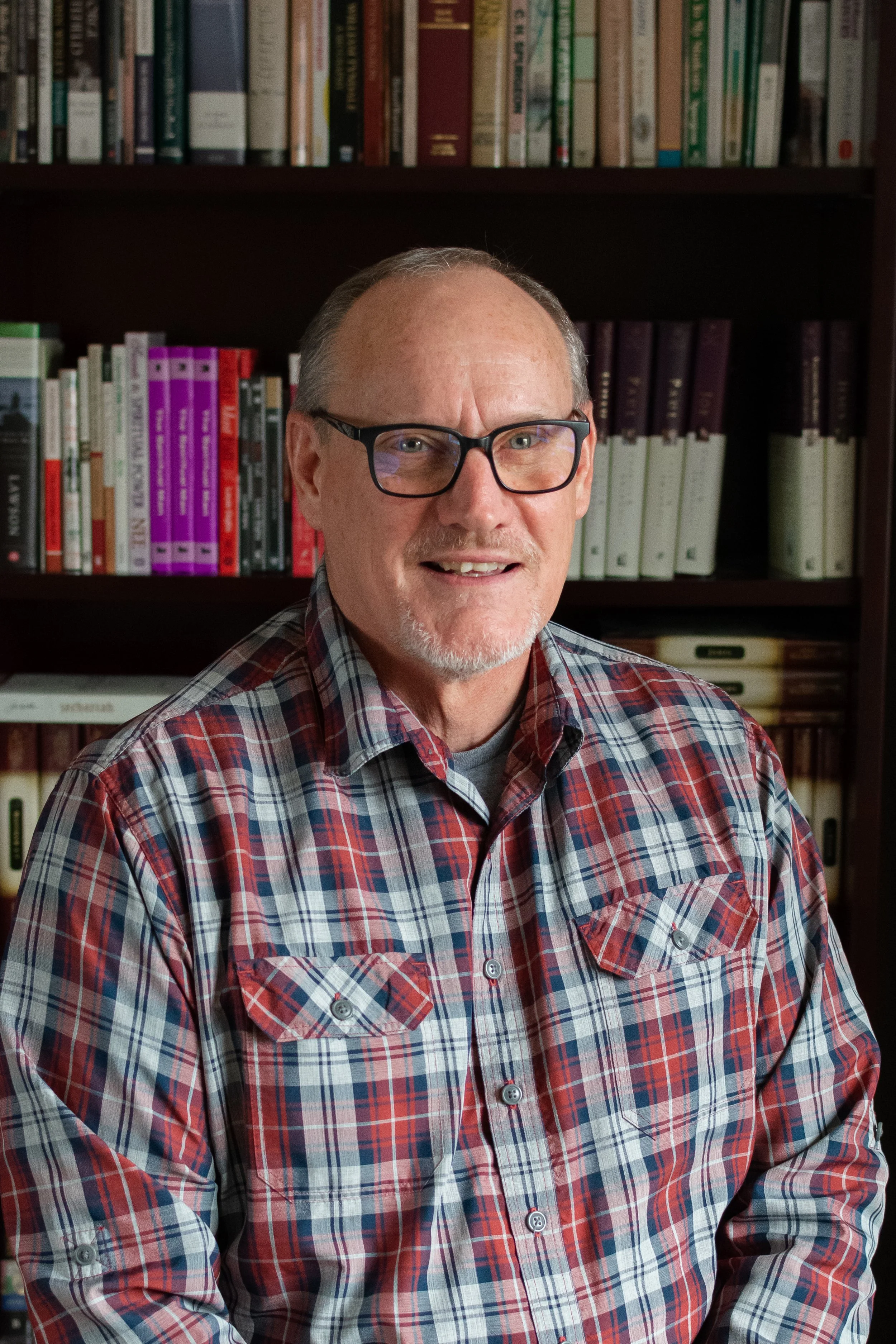 A middle-aged man with glasses, a goatee, wearing a red, gray, and blue plaid shirt standing in front of a bookshelf filled with books.