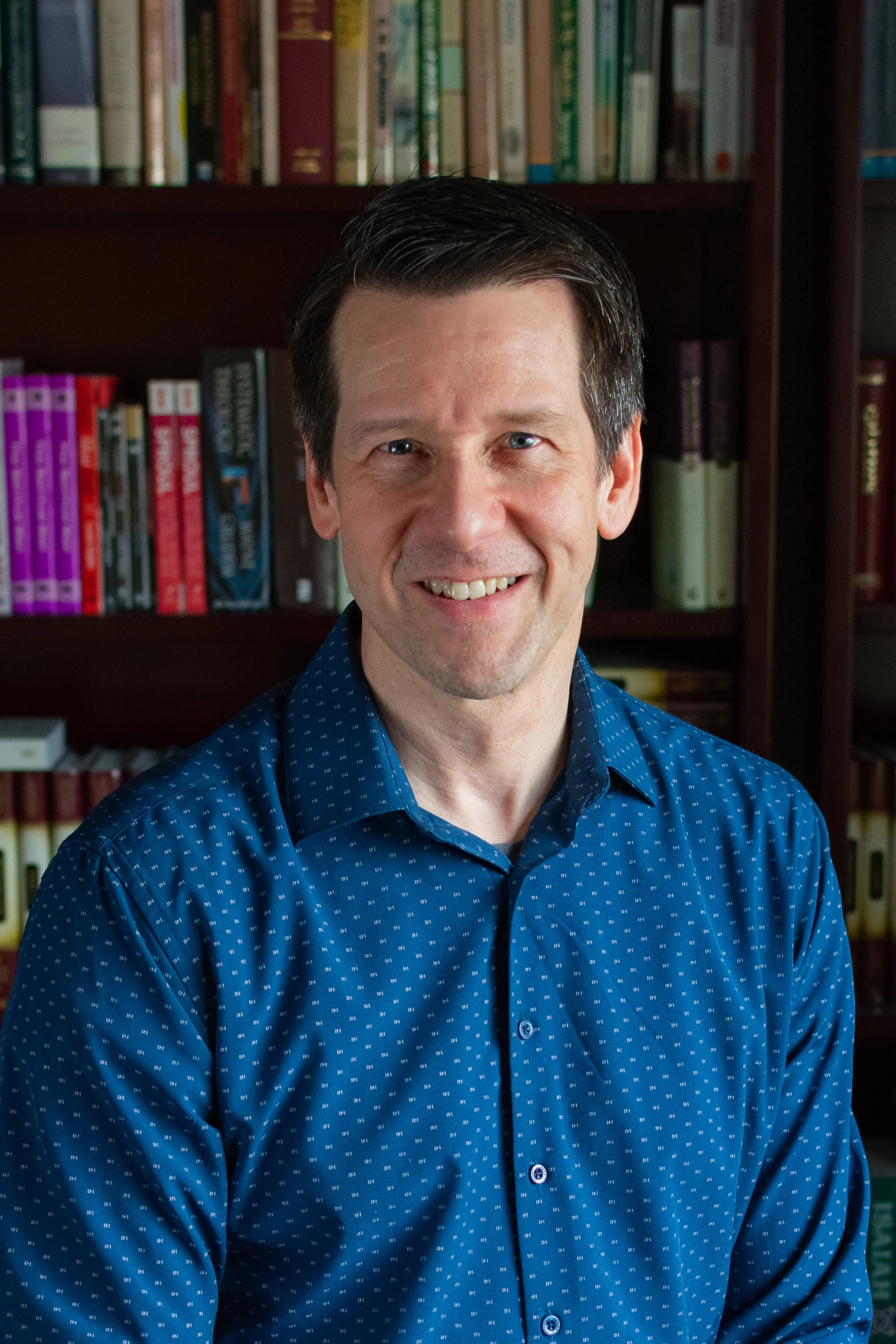 A smiling man with dark hair wearing a blue patterned shirt standing in front of a bookshelf filled with books.