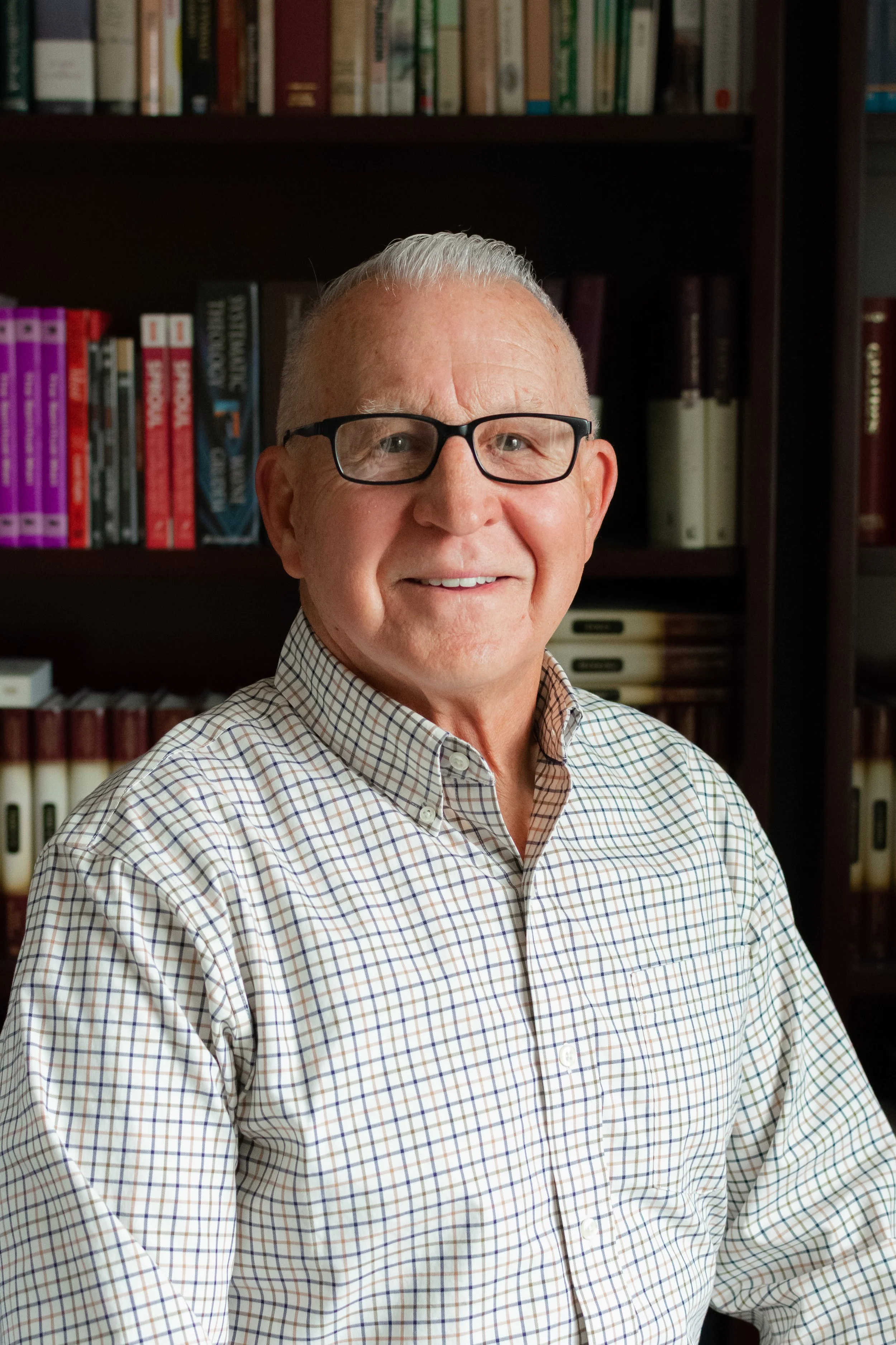 A man with gray hair, glasses, wearing a checkered shirt, standing in front of a bookshelf filled with various books.