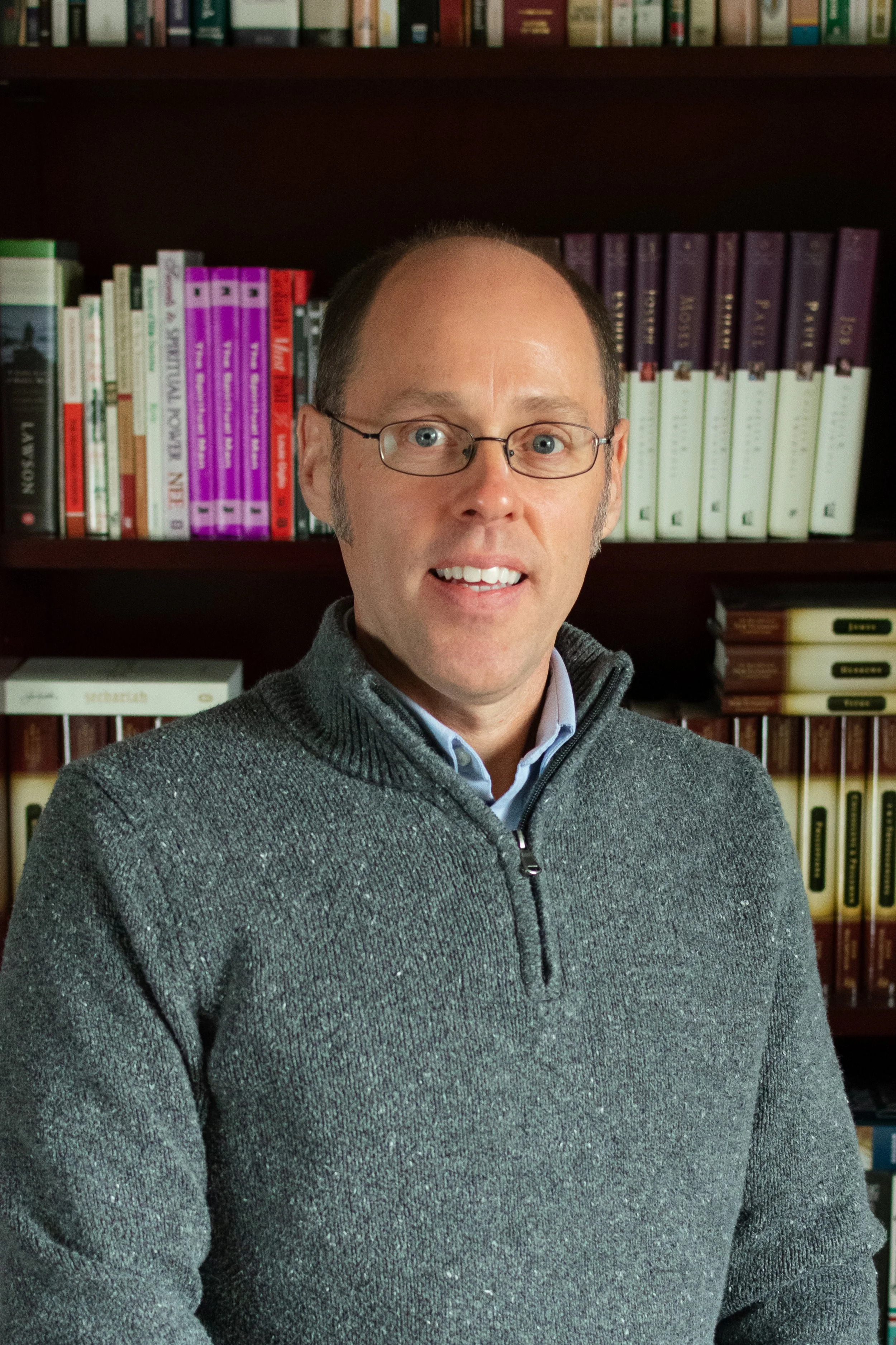 A man with glasses wearing a gray sweater, standing in front of a bookshelf filled with colorful books.