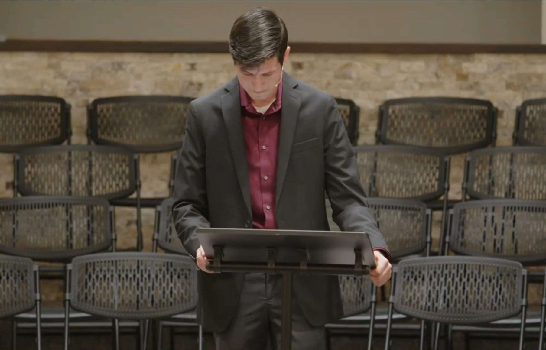 A young man with dark hair, dressed in a black suit and red shirt, looking down at a music stand in an empty room with rows of chairs and a brick wall in the background.