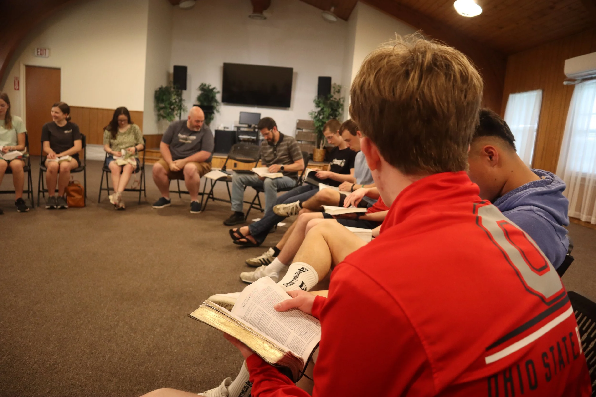 People sitting in a circle in a room with wooden walls, some with notebooks, one with a Bible, engaged in discussion or reading.