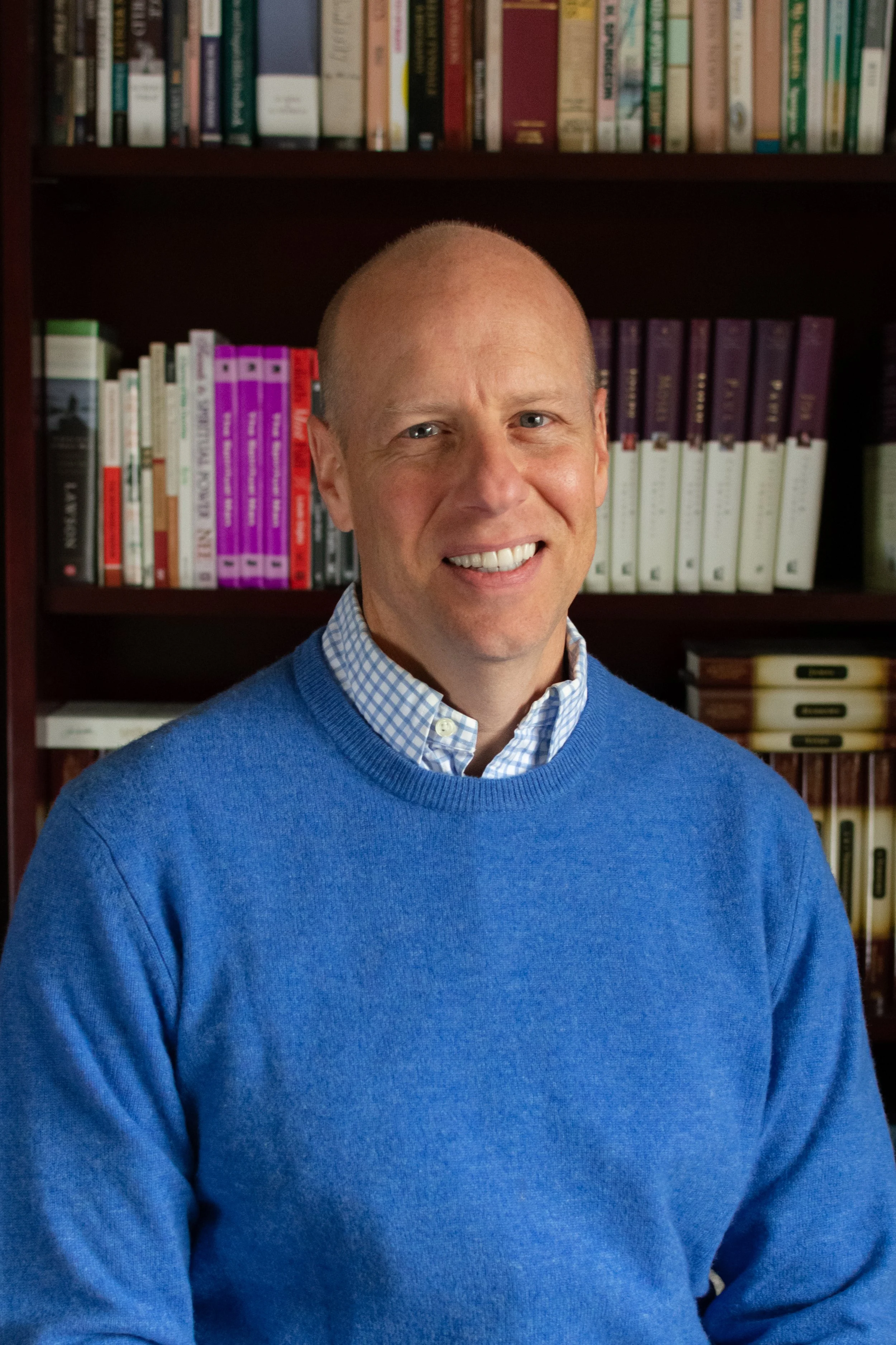 Portrait of a smiling man with a bald head, wearing a blue sweater over a checkered collared shirt. He is standing in front of a bookshelf filled with books.