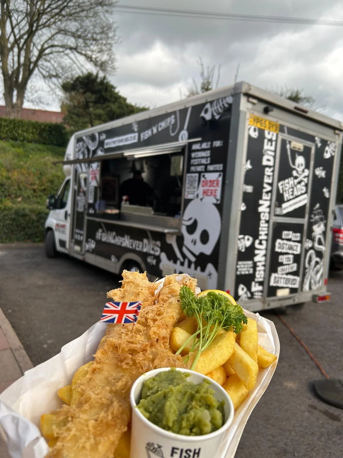 Fried fish fillets with lemon, garnished with parsley, served with French fries, a small cup of green sauce, and a side of lemon wedges in a food tray, with a fish and chips food truck in the background.