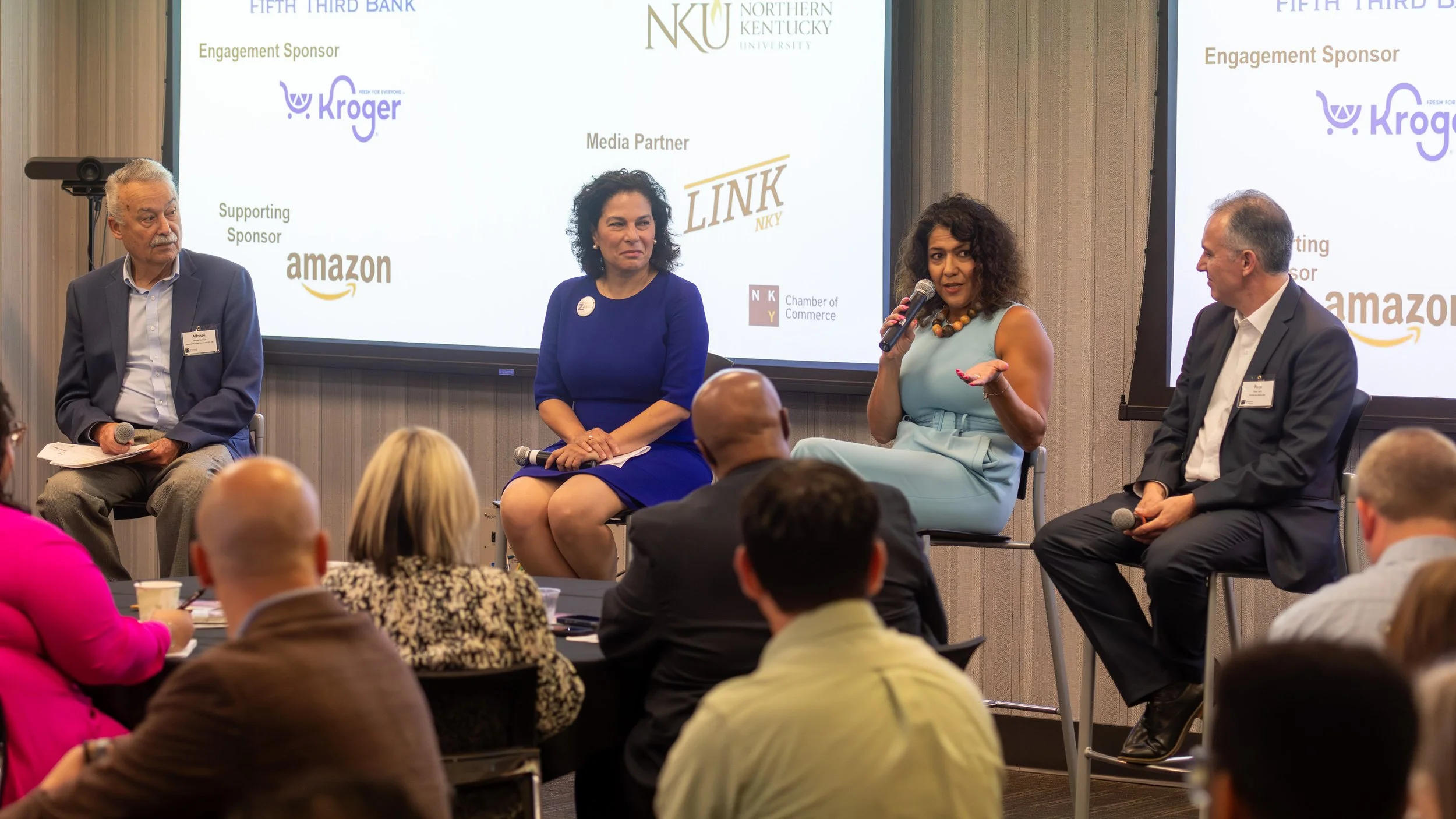 A panel discussion with four speakers sitting on stage, two men and two women. One woman is speaking into a microphone while gesturing with her hand. The background has a large screen displaying logos of NKU, Kroger, Amazon, LINK Magazine, and the Ch